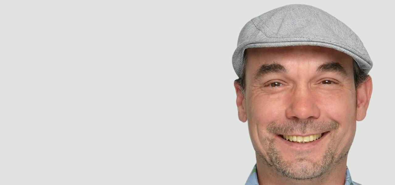 Lächelnder Mann mit grauer Schiebermütze und leichtem Bart vor hellem Hintergrund.
