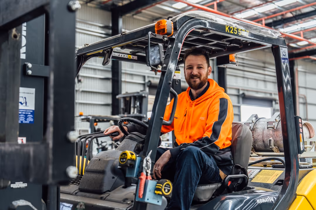 Smiling man in an orange hoodie sitting in the driver's seat of a forklift in a warehouse.