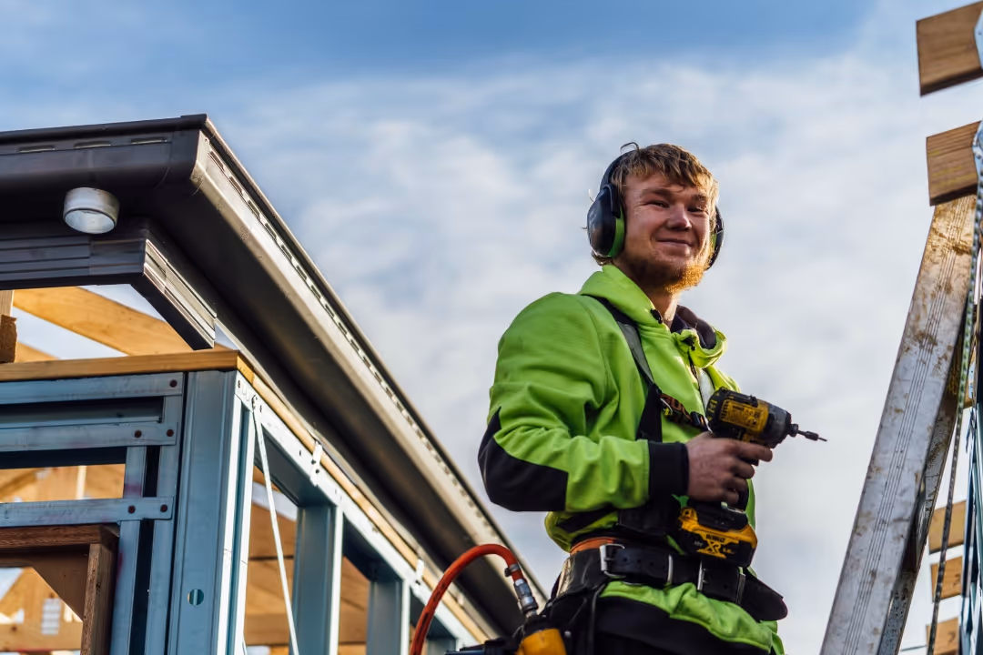 Construction worker in green jacket using a power drill on a building frame outdoors, smiling.