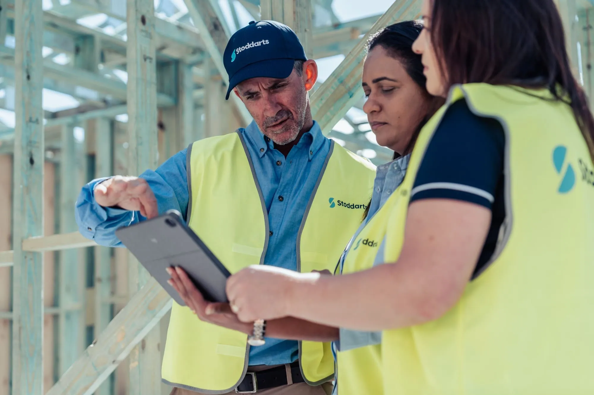 Three construction workers in yellow vests looking at a tablet inside a wooden framework building.