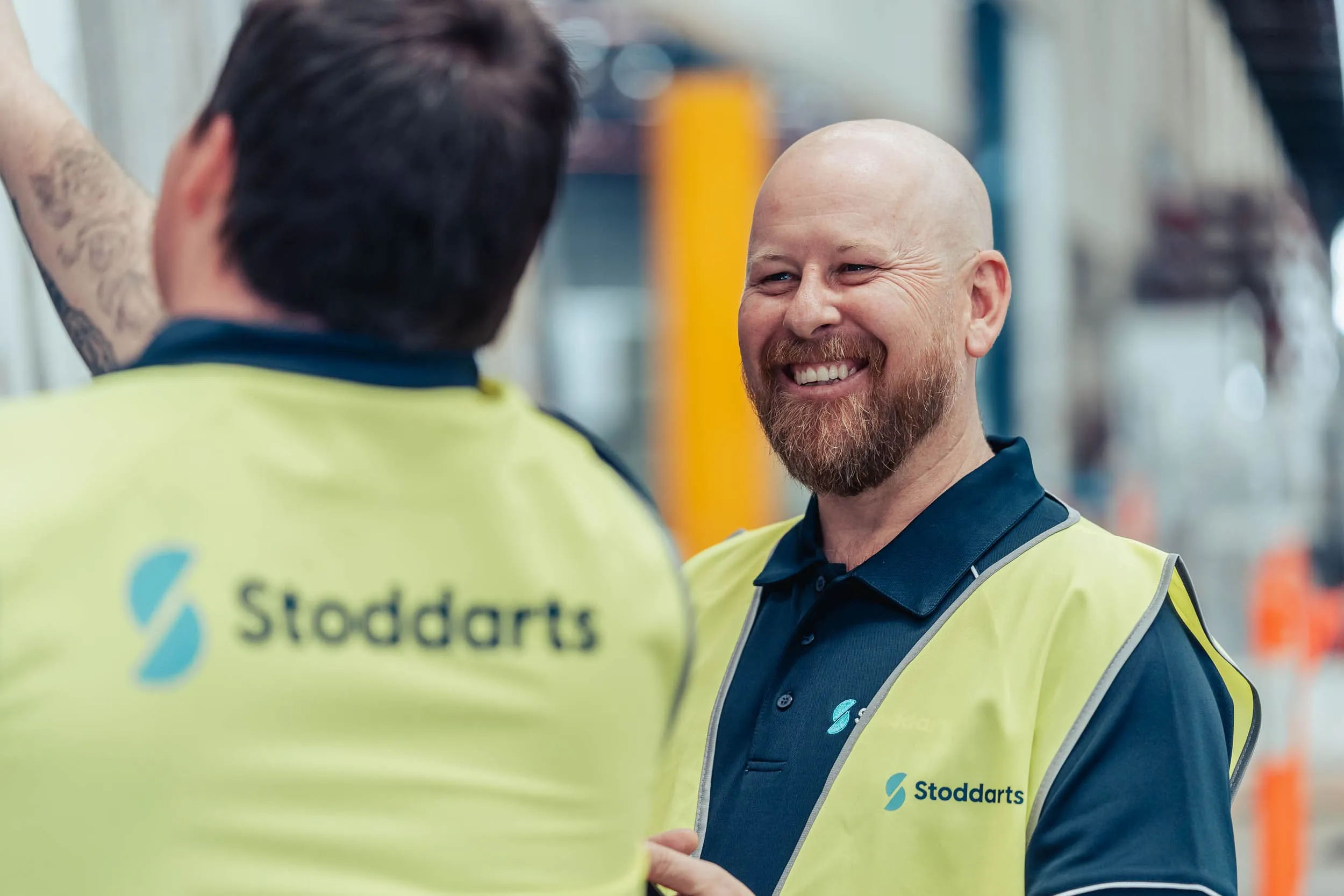 Two men in Stoddarts safety vests talking, one smiling and the other raising his arm indoors.