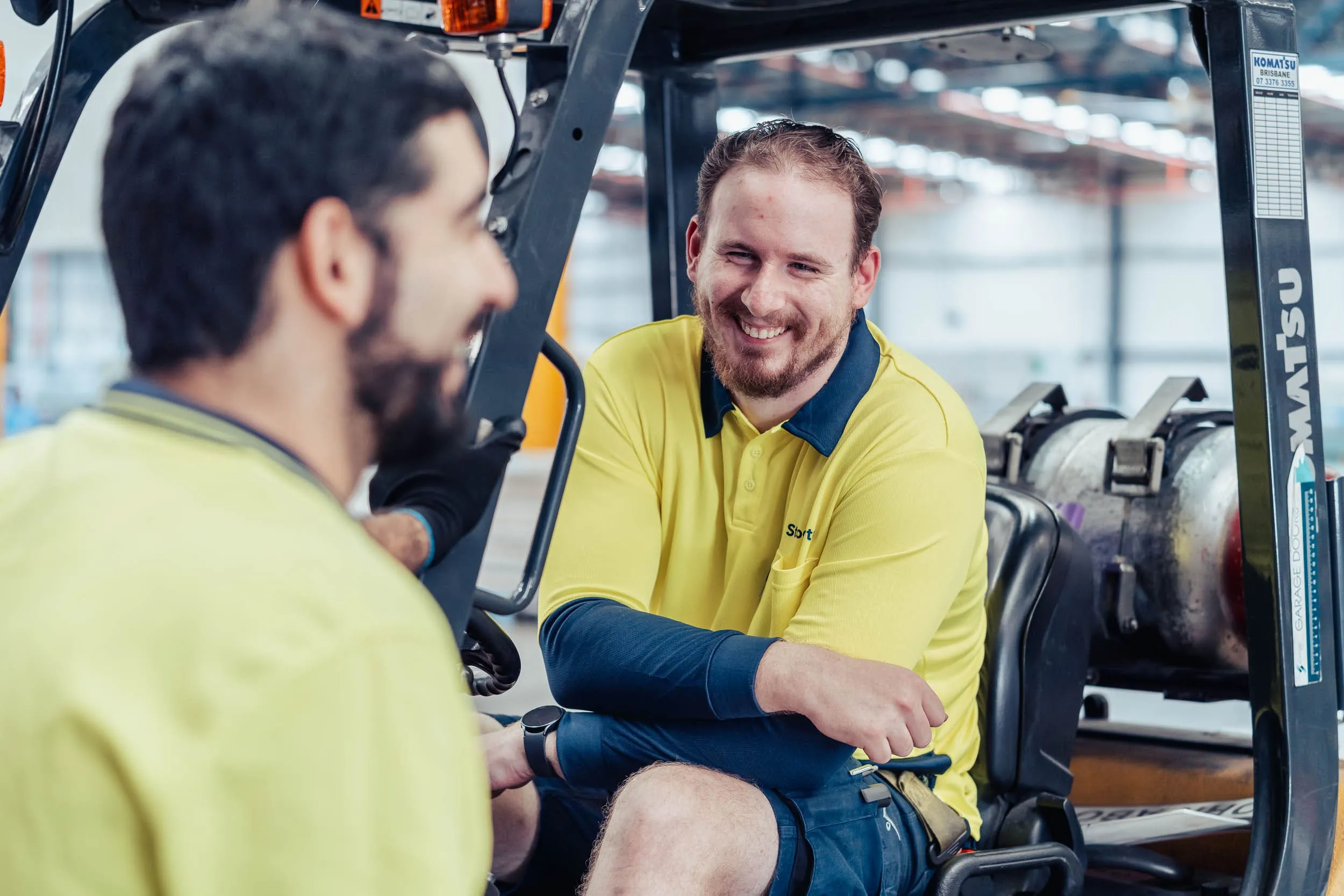 Two male warehouse workers in yellow shirts smiling and chatting near a forklift.