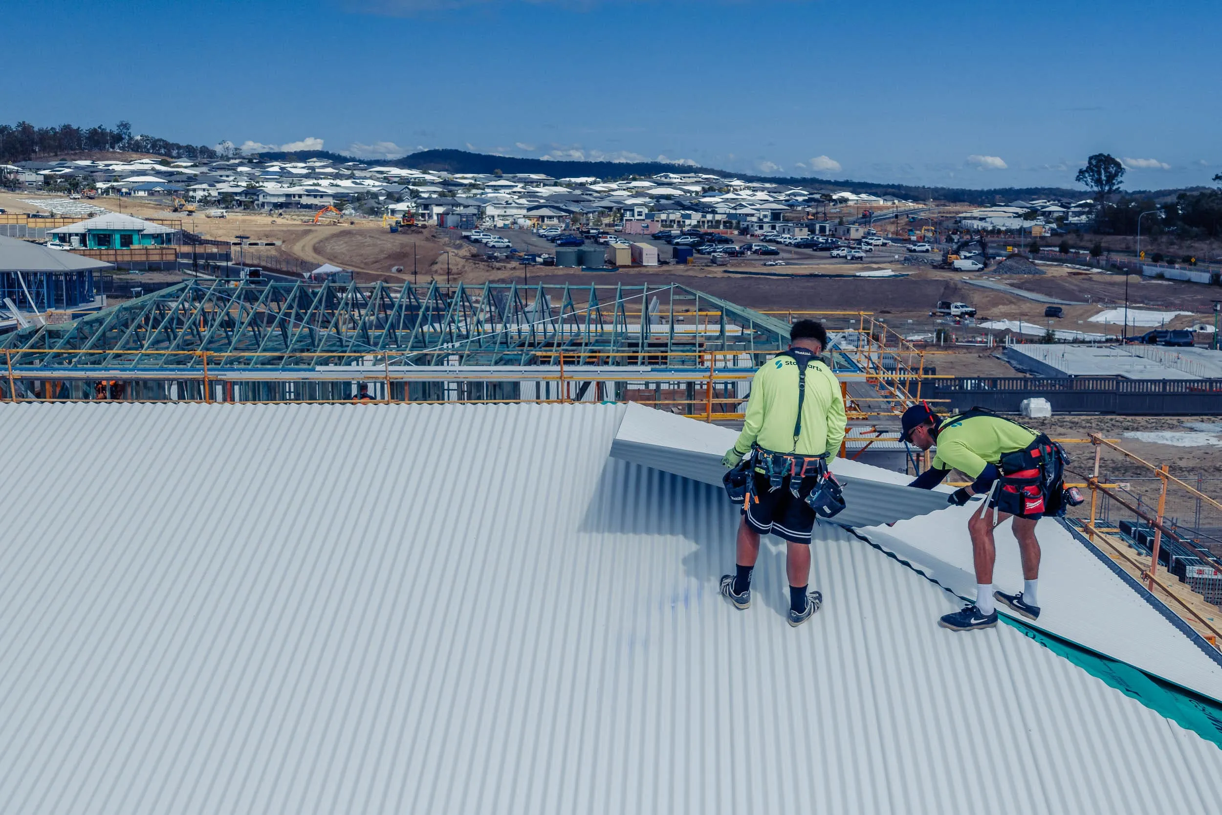 Two construction workers installing large corrugated metal roofing panels on a building under construction.