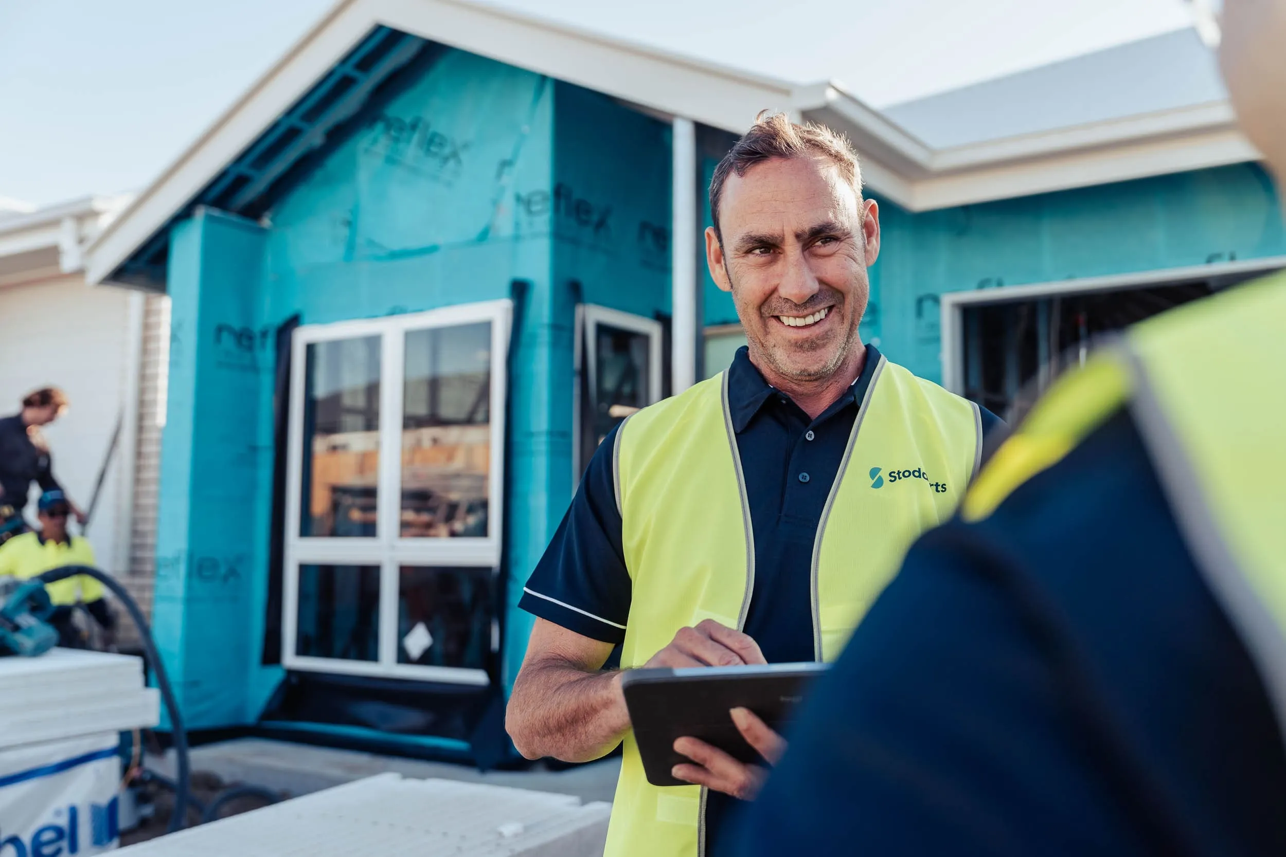 Smiling construction worker wearing a yellow vest using a tablet in front of a house under construction.