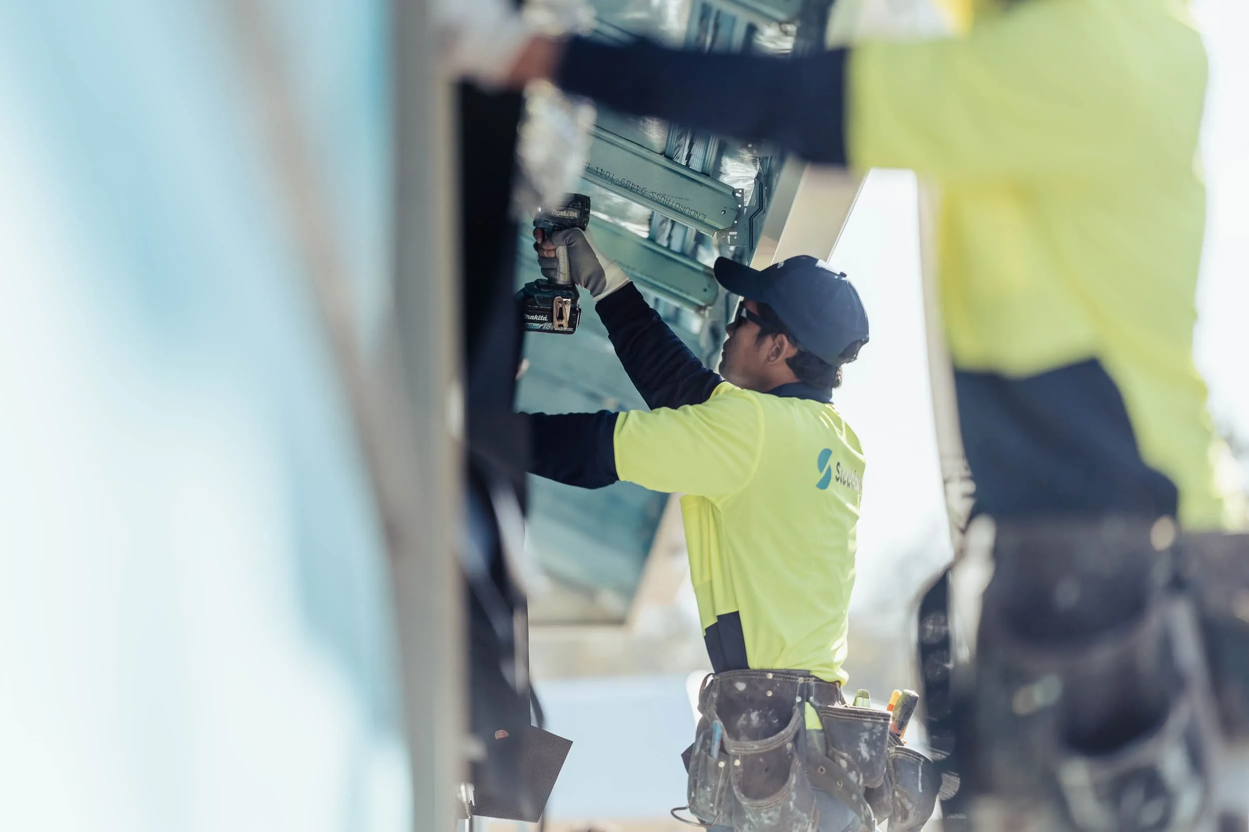 Construction worker in yellow shirt using a power drill on metal framing, wearing a tool belt.