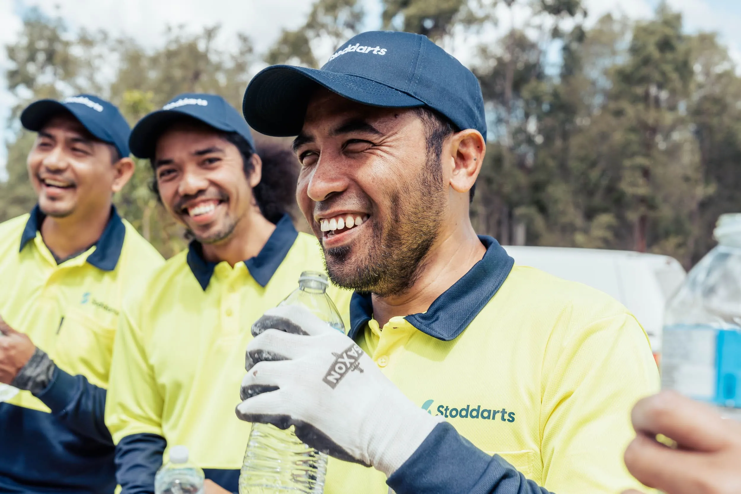 Three men in yellow Stoddarts uniforms and caps smiling and holding water bottles outdoors.