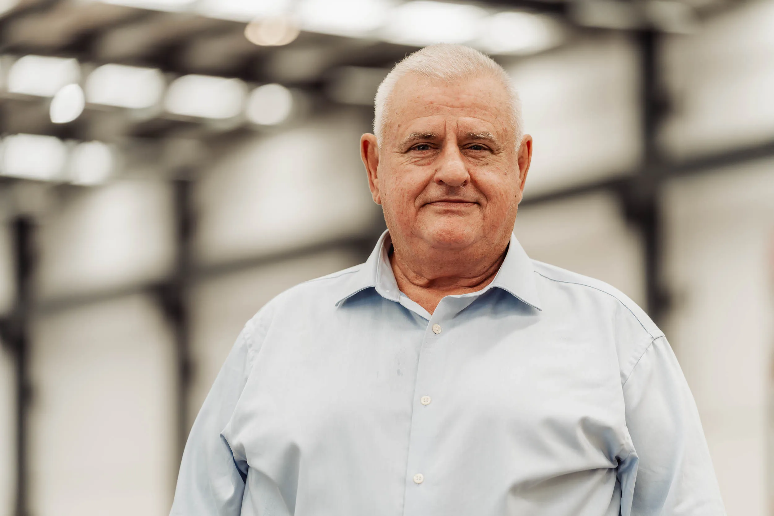 Older man with short gray hair wearing a light blue button-up shirt, standing inside a large building.