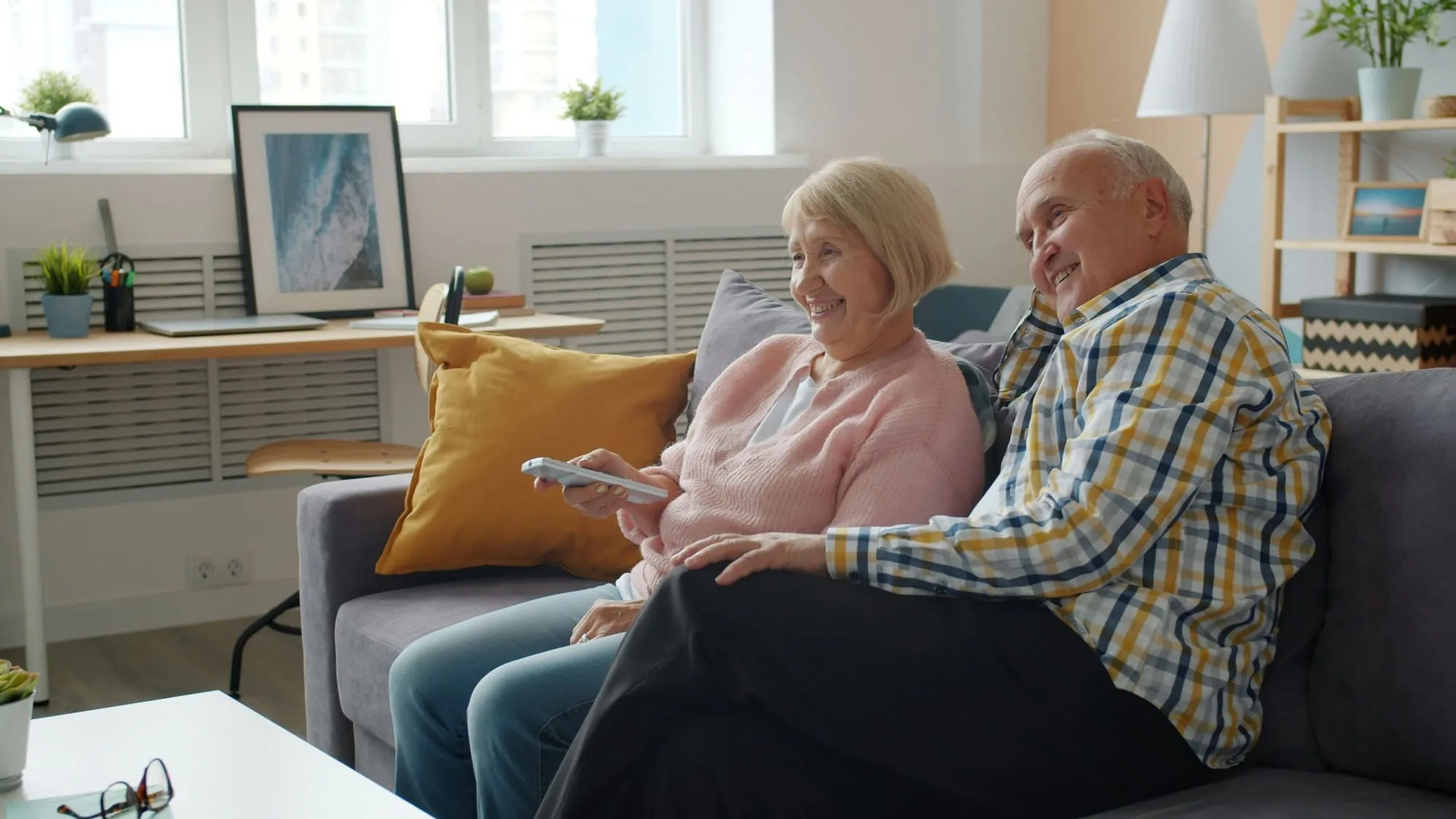 Smiling elderly couple sitting on couch watching TV with remote control in hand in cozy living room.