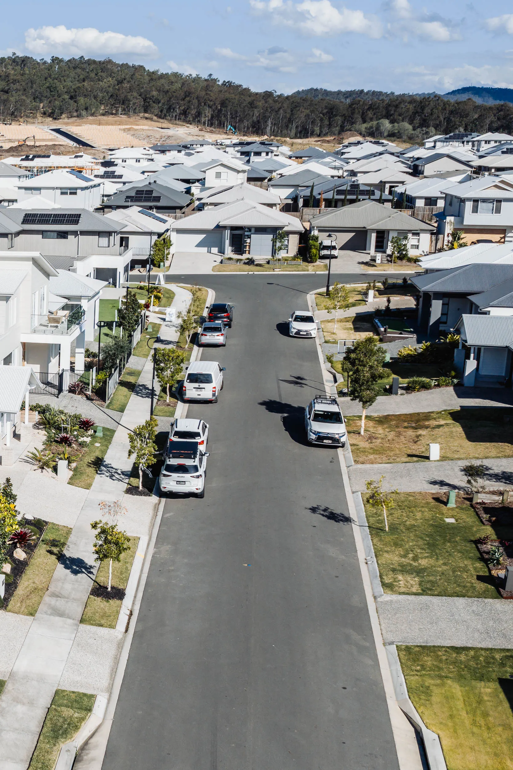Residential street with parked cars and modern houses having gray or white roofs under a blue sky.