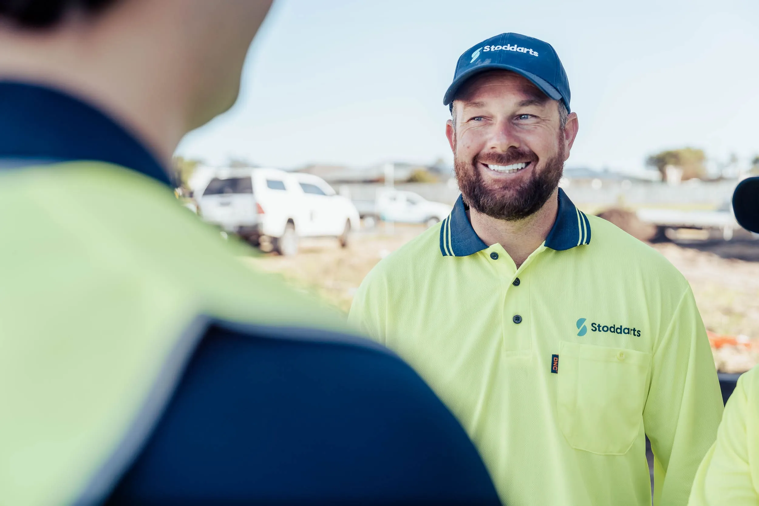 Smiling man wearing a Stoddarts cap and high-visibility shirt talking to a colleague outdoors.