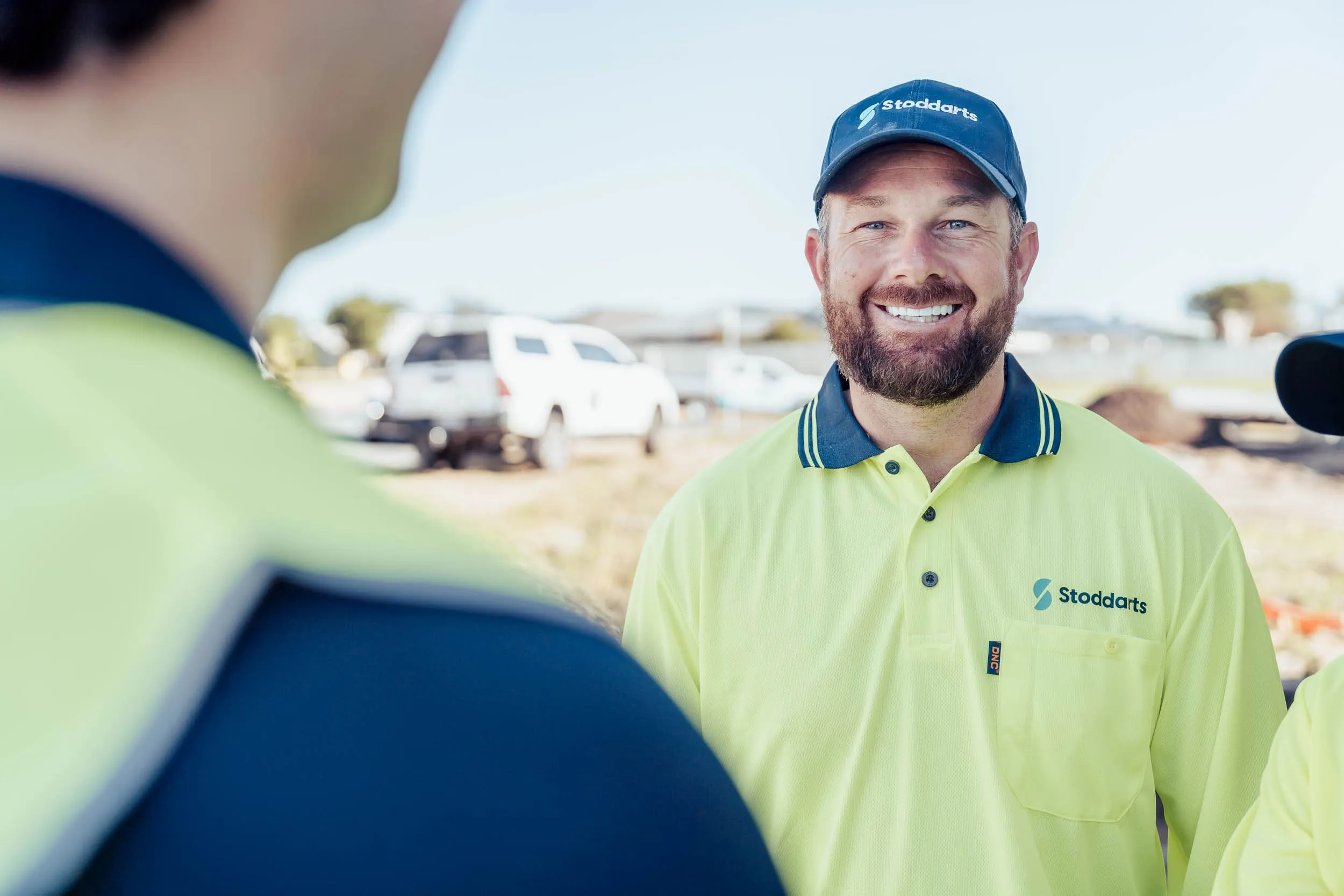 Smiling man in a yellow Stoddarts uniform and cap on a construction site with blurred people and vehicles.