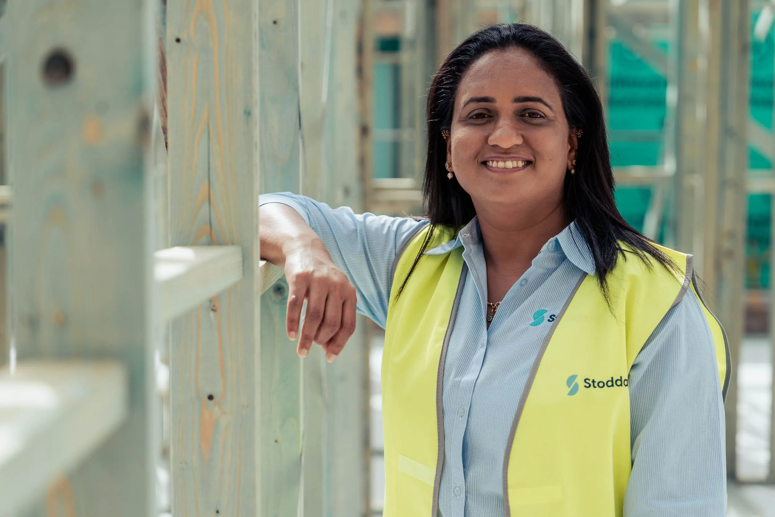 Smiling woman in a high-visibility vest leaning on wood framing at a construction site.