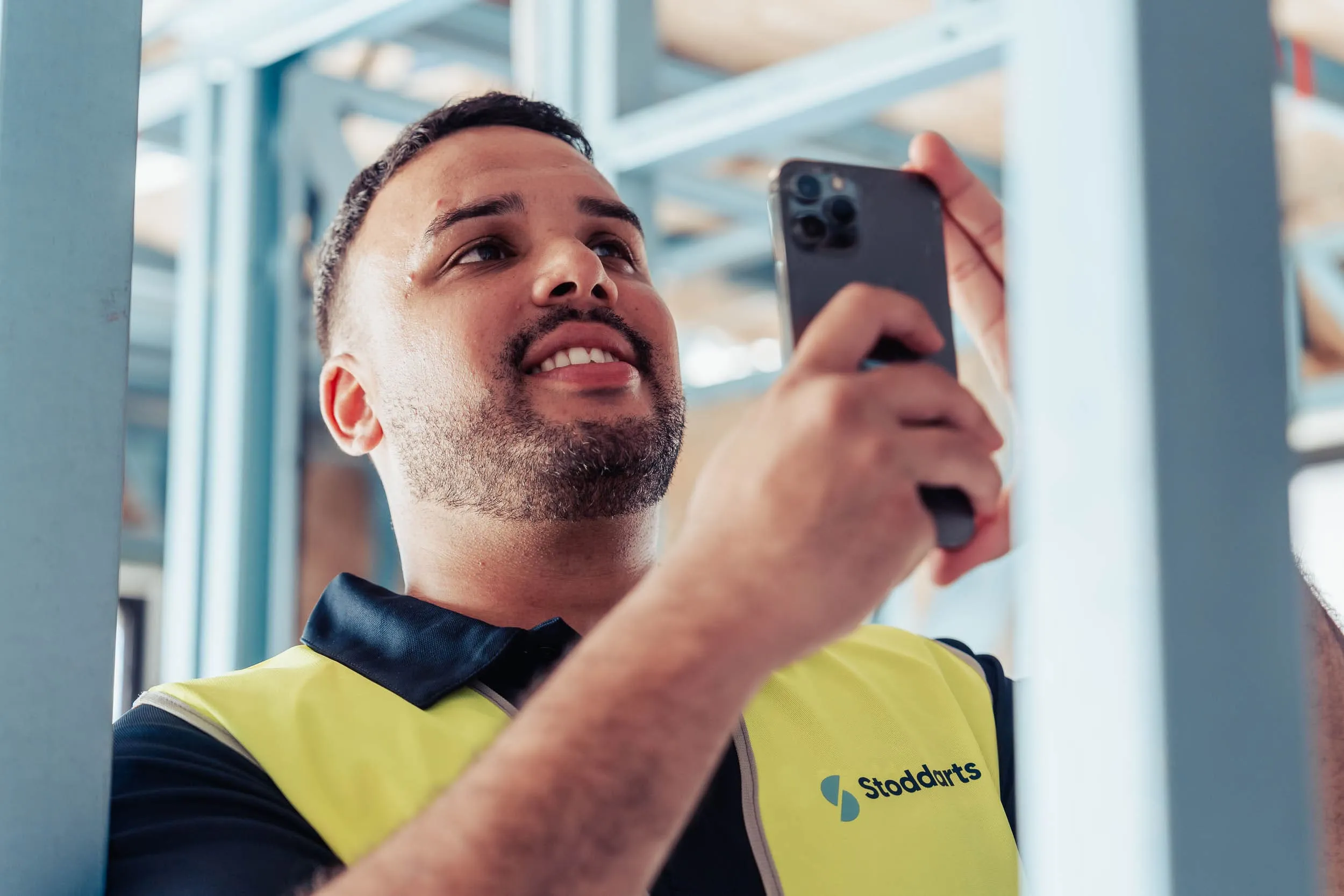 Man in a Stoddarts work vest holding a smartphone, taking a photo or video indoors.