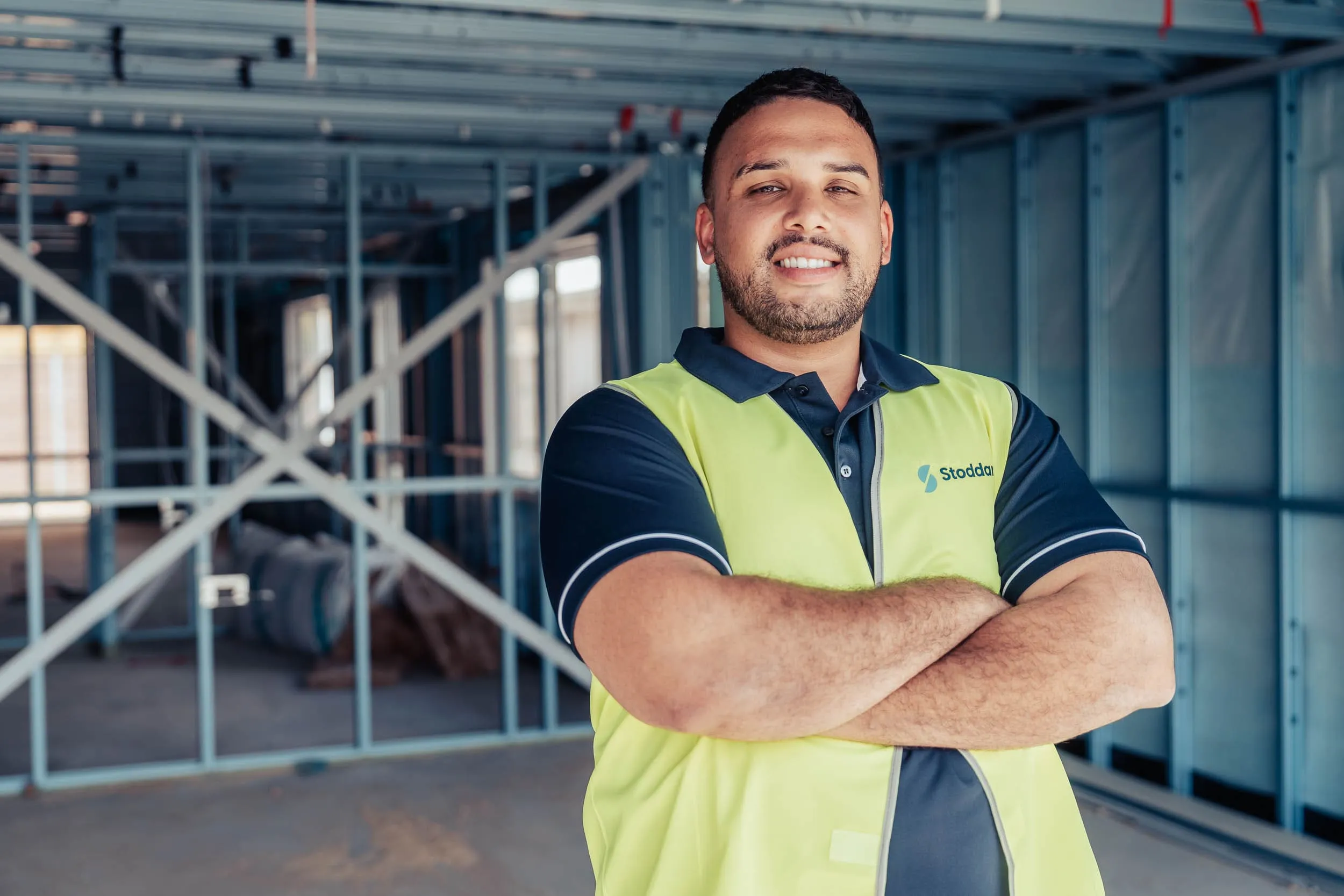 Confident construction worker with crossed arms wearing a yellow safety vest and navy shirt inside a building site.