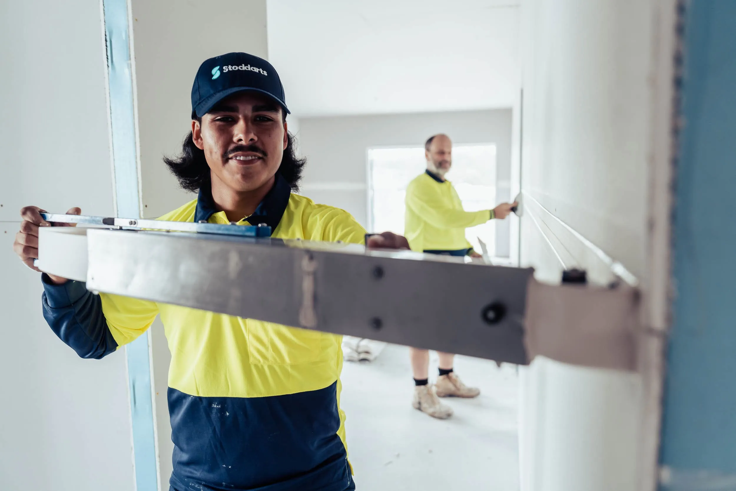 Two construction workers in yellow shirts using a level tool on a white wall indoors.
