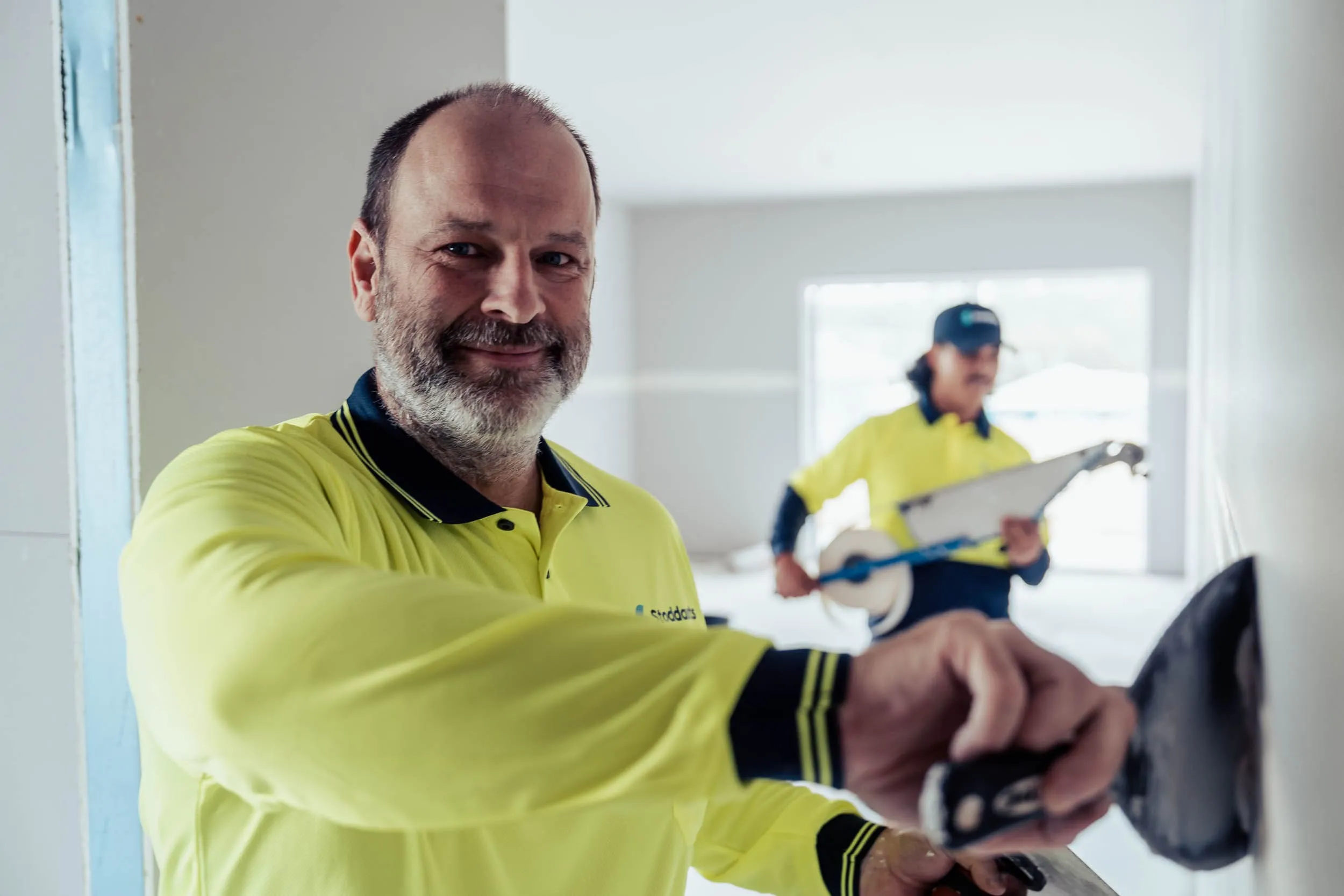 Two construction workers in yellow shirts working inside a bright room, one smoothing a wall.