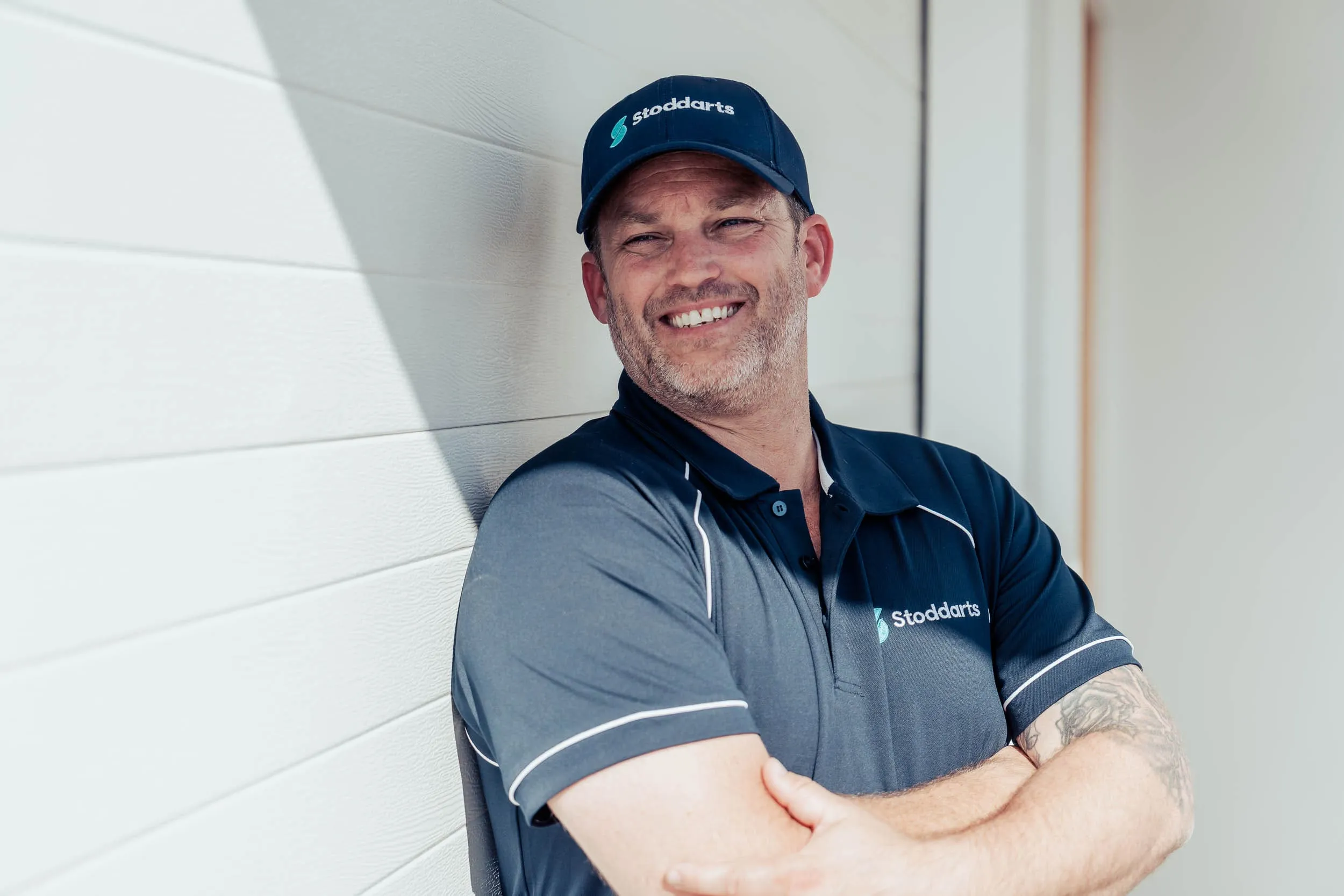 Smiling man with a beard wearing a navy blue Stoddarts hat and shirt, leaning against a white wall.