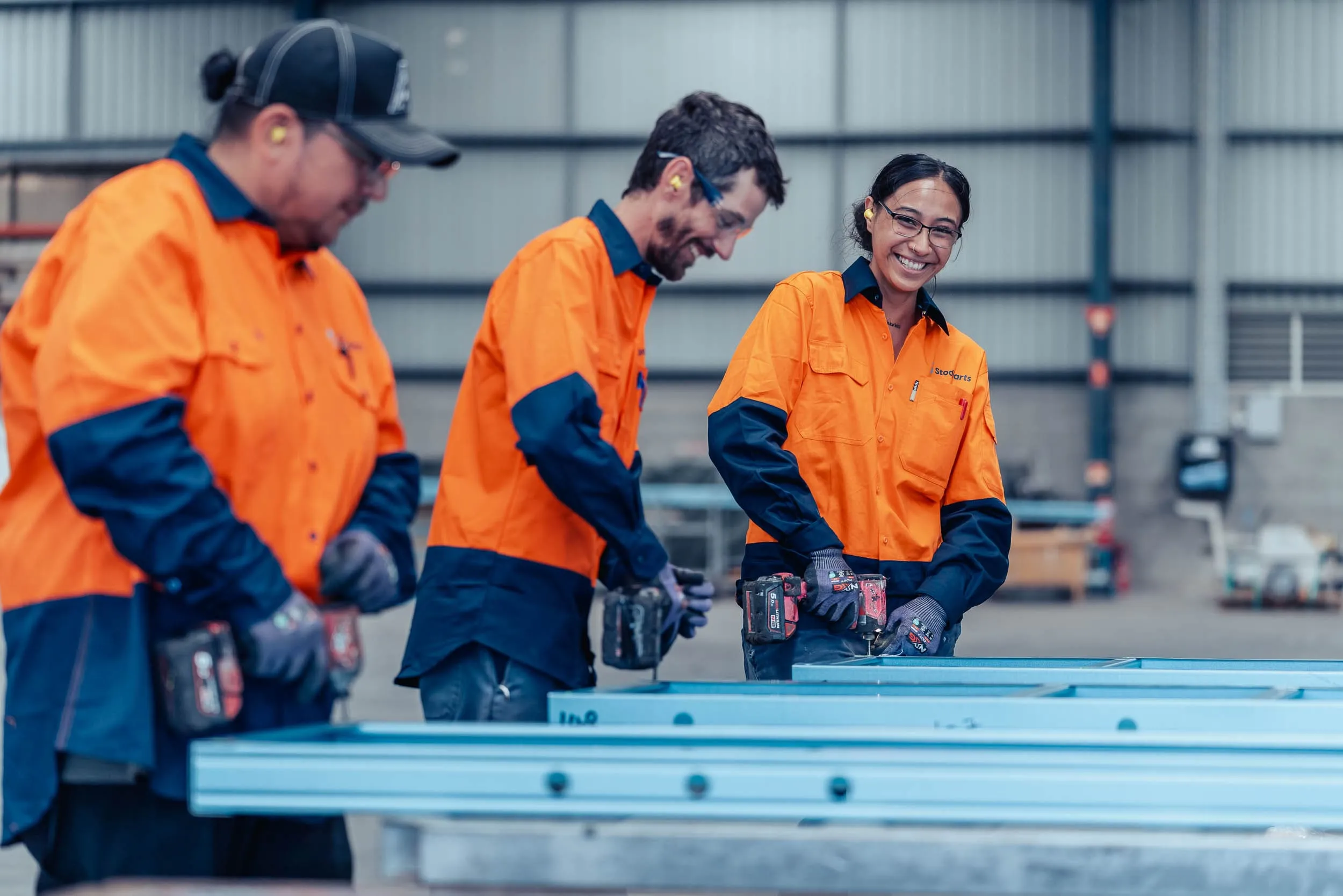 Three workers in orange and navy uniforms using power drills on metal frames in a warehouse.