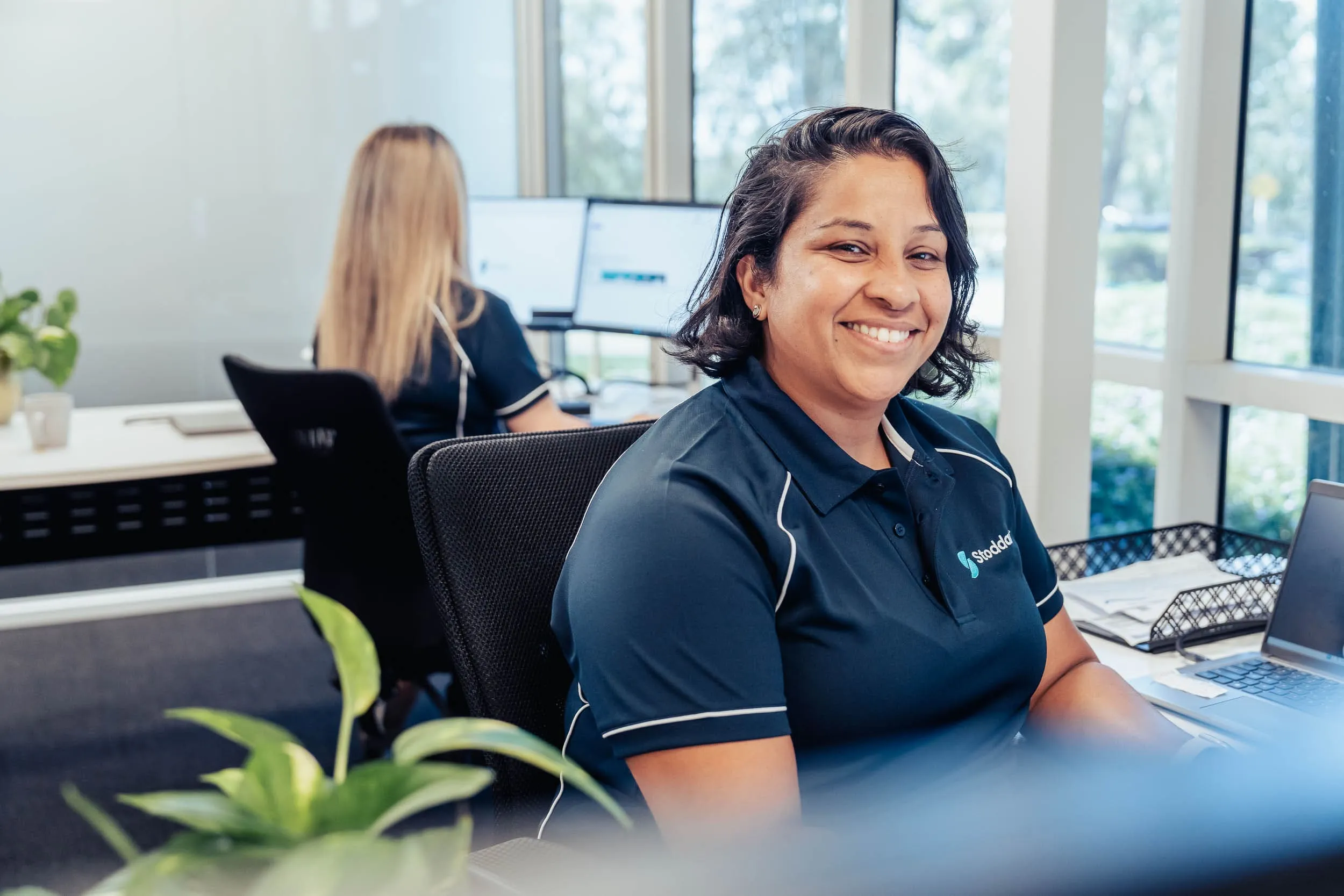 Smiling woman in a navy polo shirt sitting at a desk in an office with another woman working at computers.