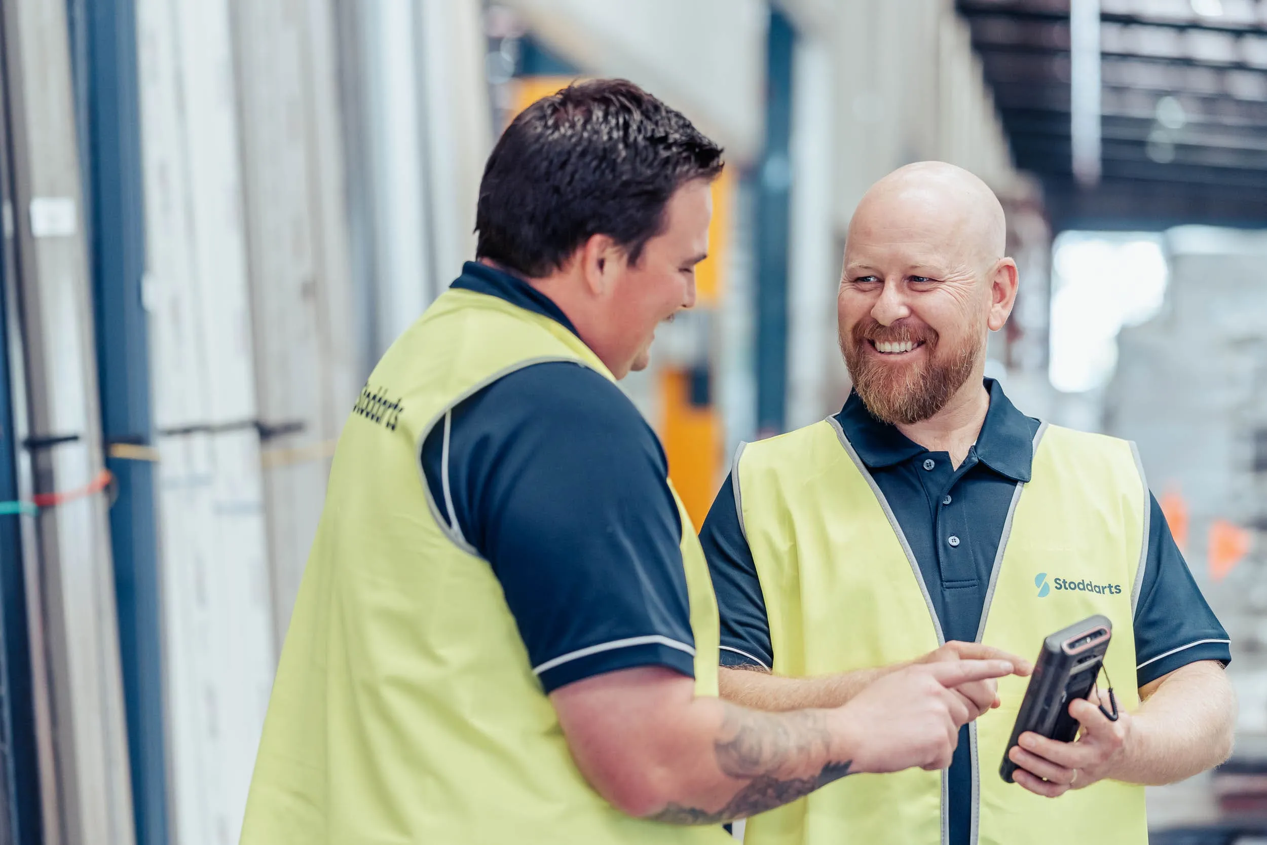 Two men in yellow vests smiling and using a handheld device inside a warehouse.
