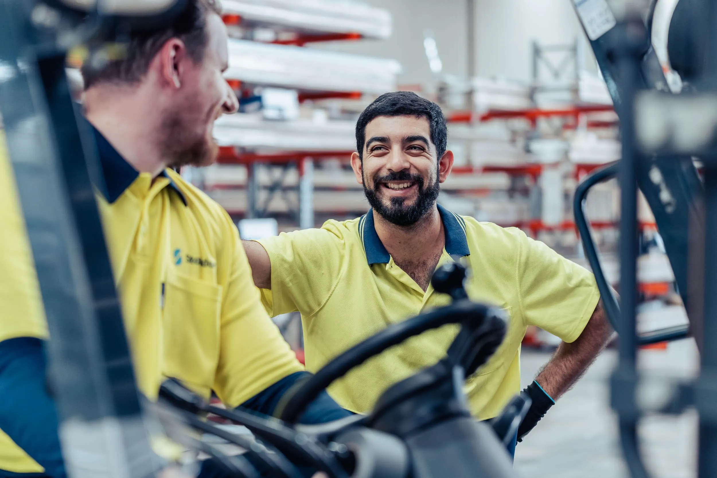 Two warehouse workers in yellow shirts smiling and chatting near forklift trucks.