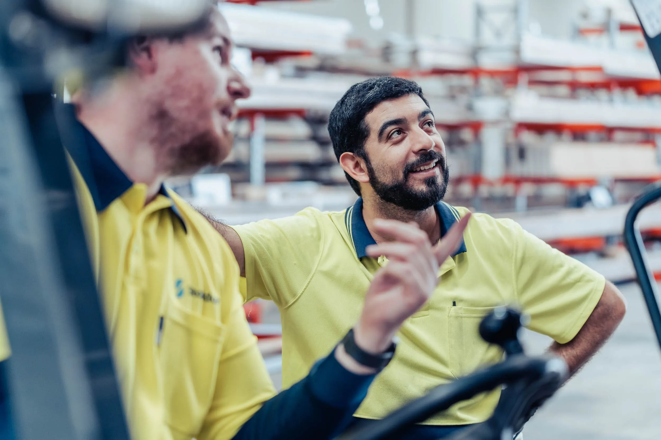 Two warehouse workers in yellow shirts engaged in conversation with shelves in the background.