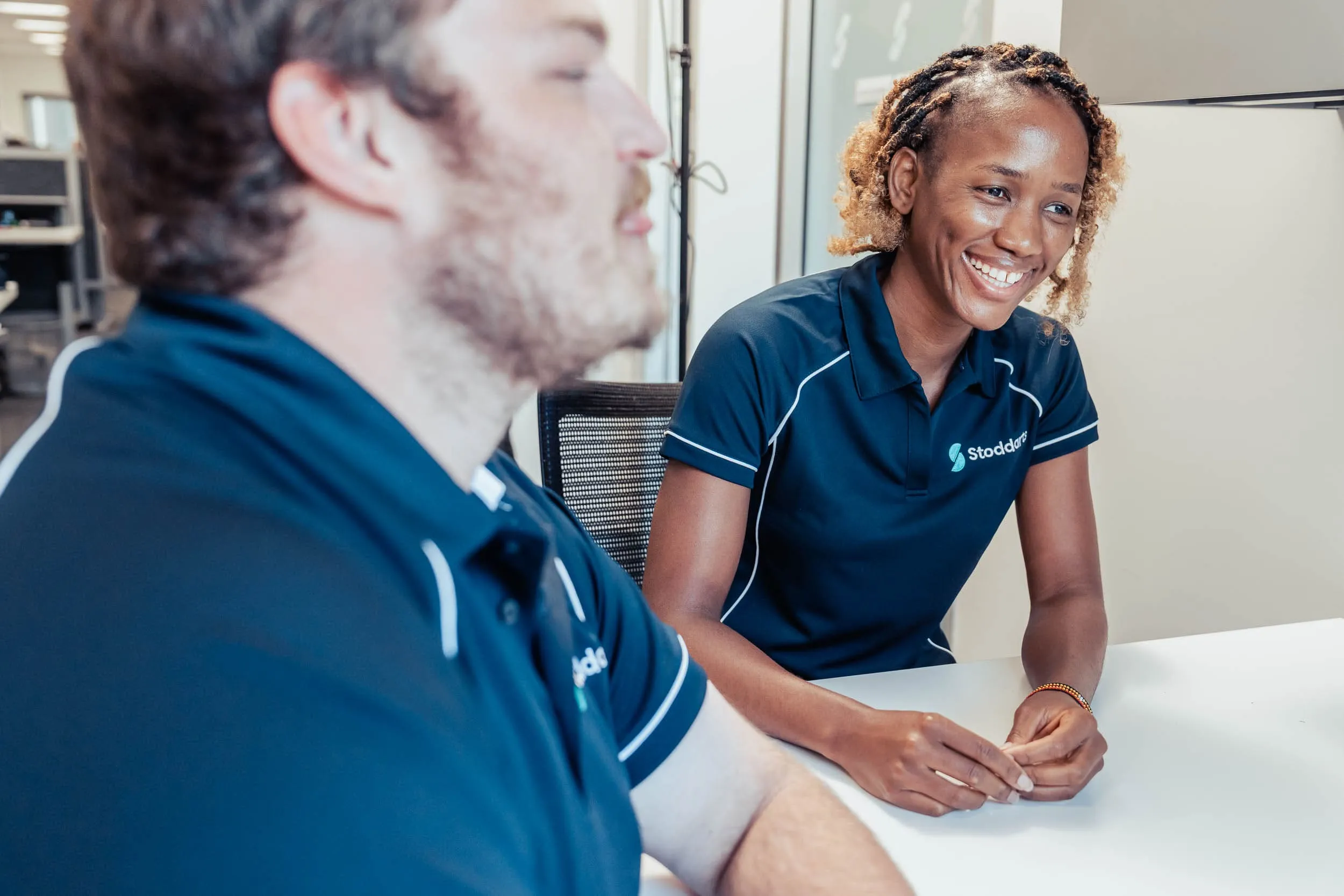 Two colleagues in navy Stoddart polo shirts having a friendly conversation at a white table.