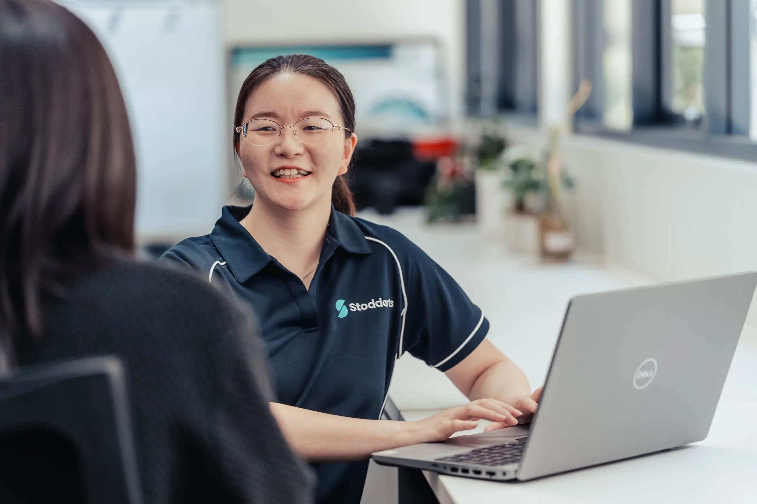 Smiling woman wearing glasses and a Stoddarts polo shirt typing on a Dell laptop across from another person.