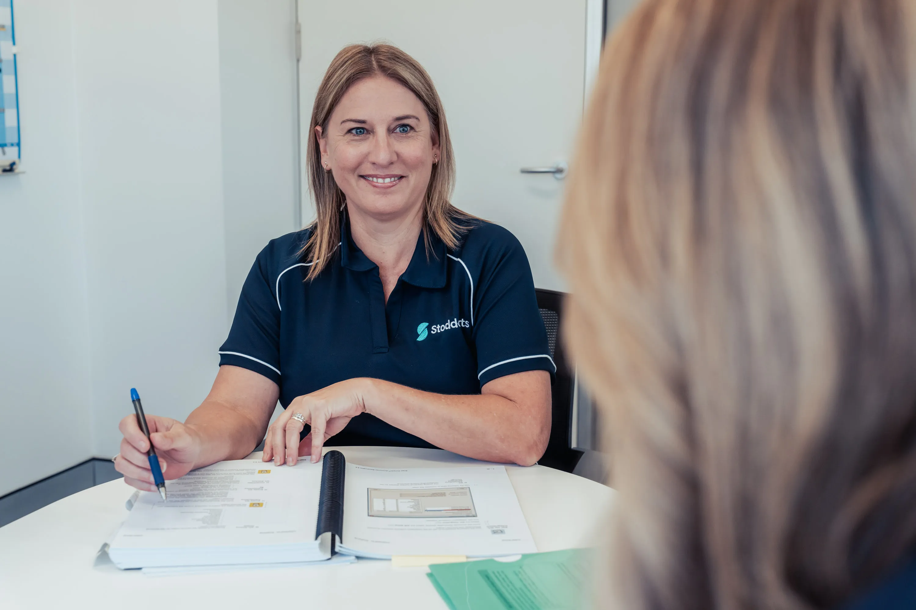 Woman in blue Stoddarts shirt smiling and holding a pen over open documents at a white table.
