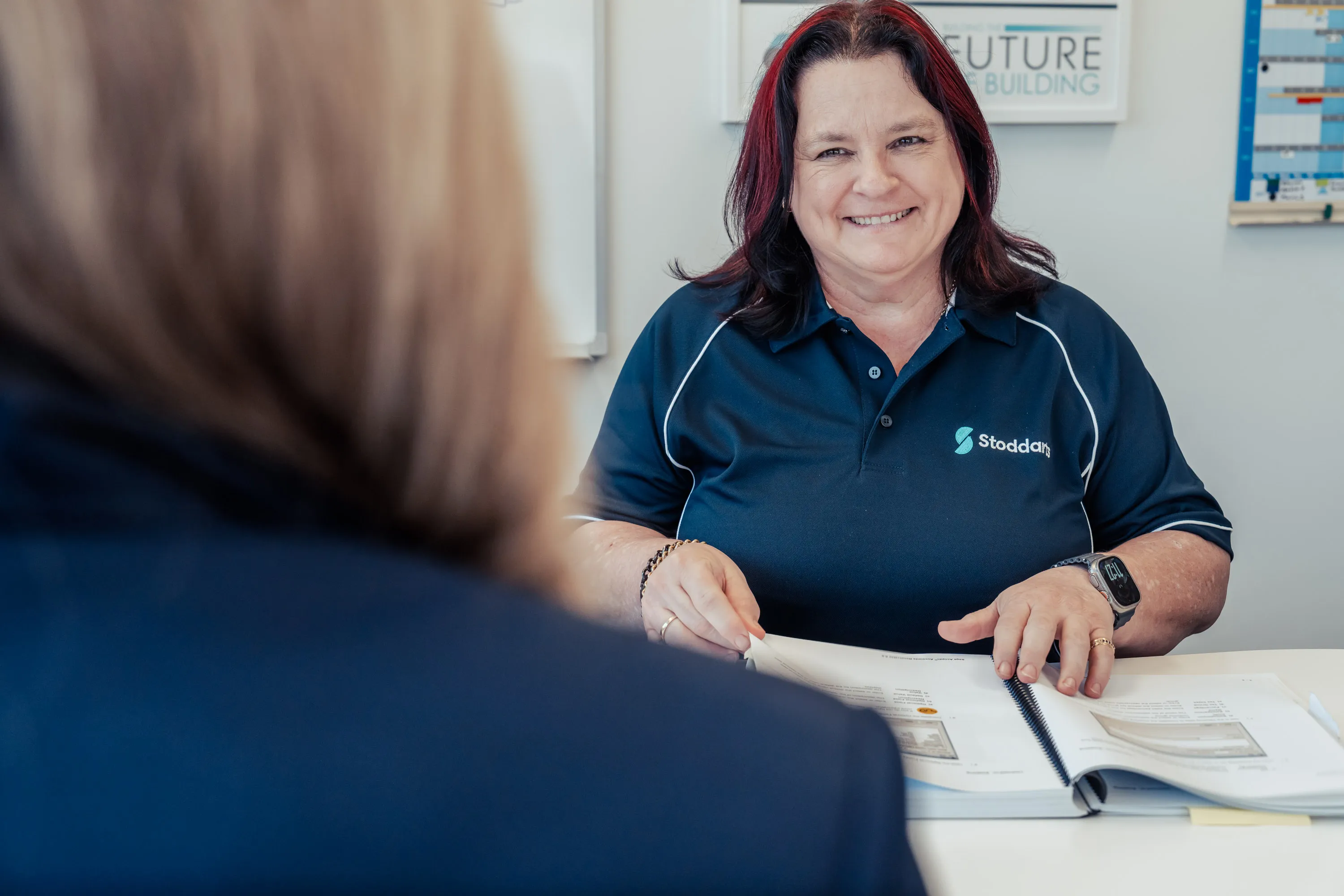 Smiling woman with red highlights in a Stoddarts shirt showing a spiral-bound booklet to another person.