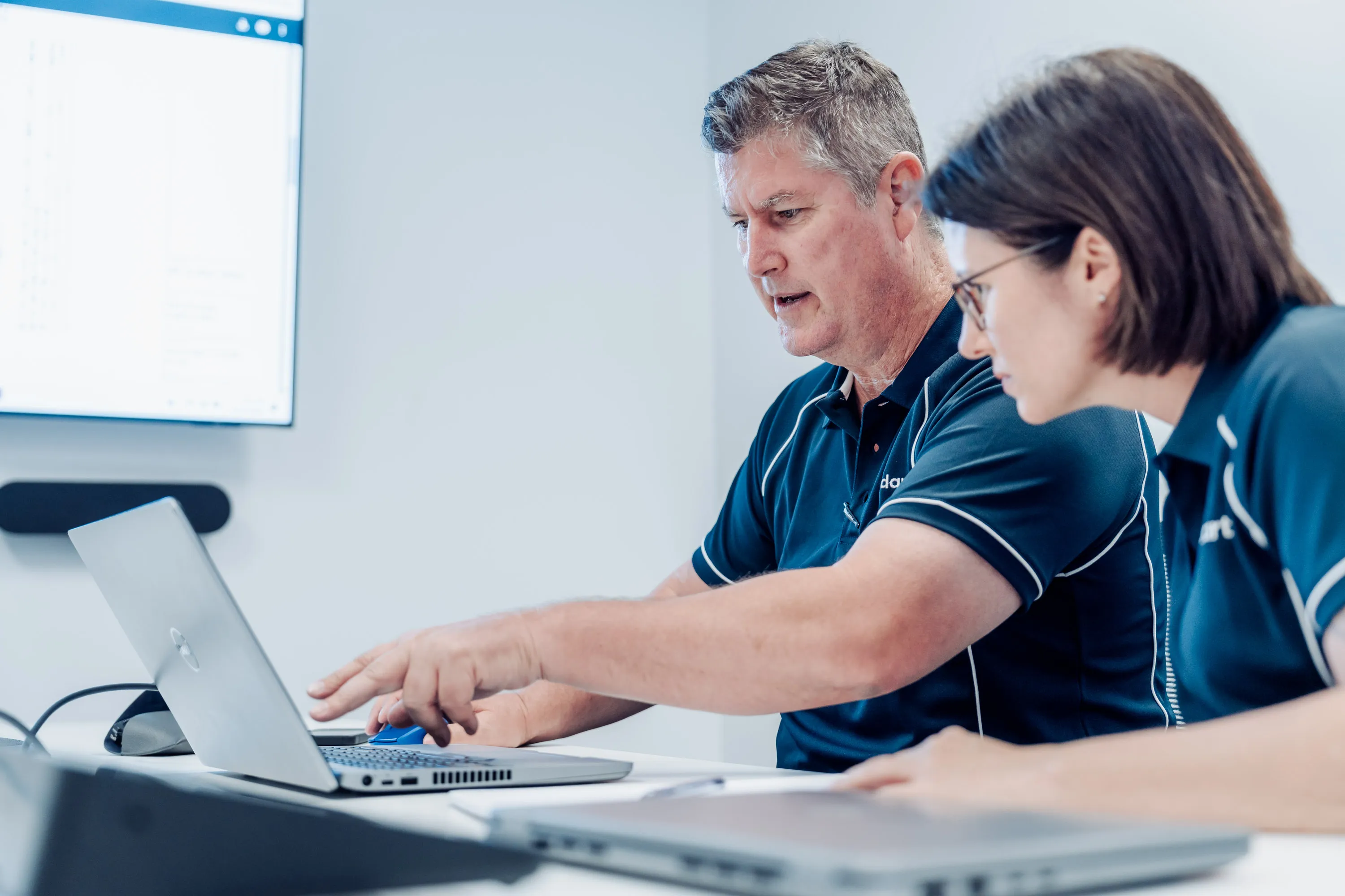Two people in blue shirts looking at and pointing to a laptop screen in an office setting.