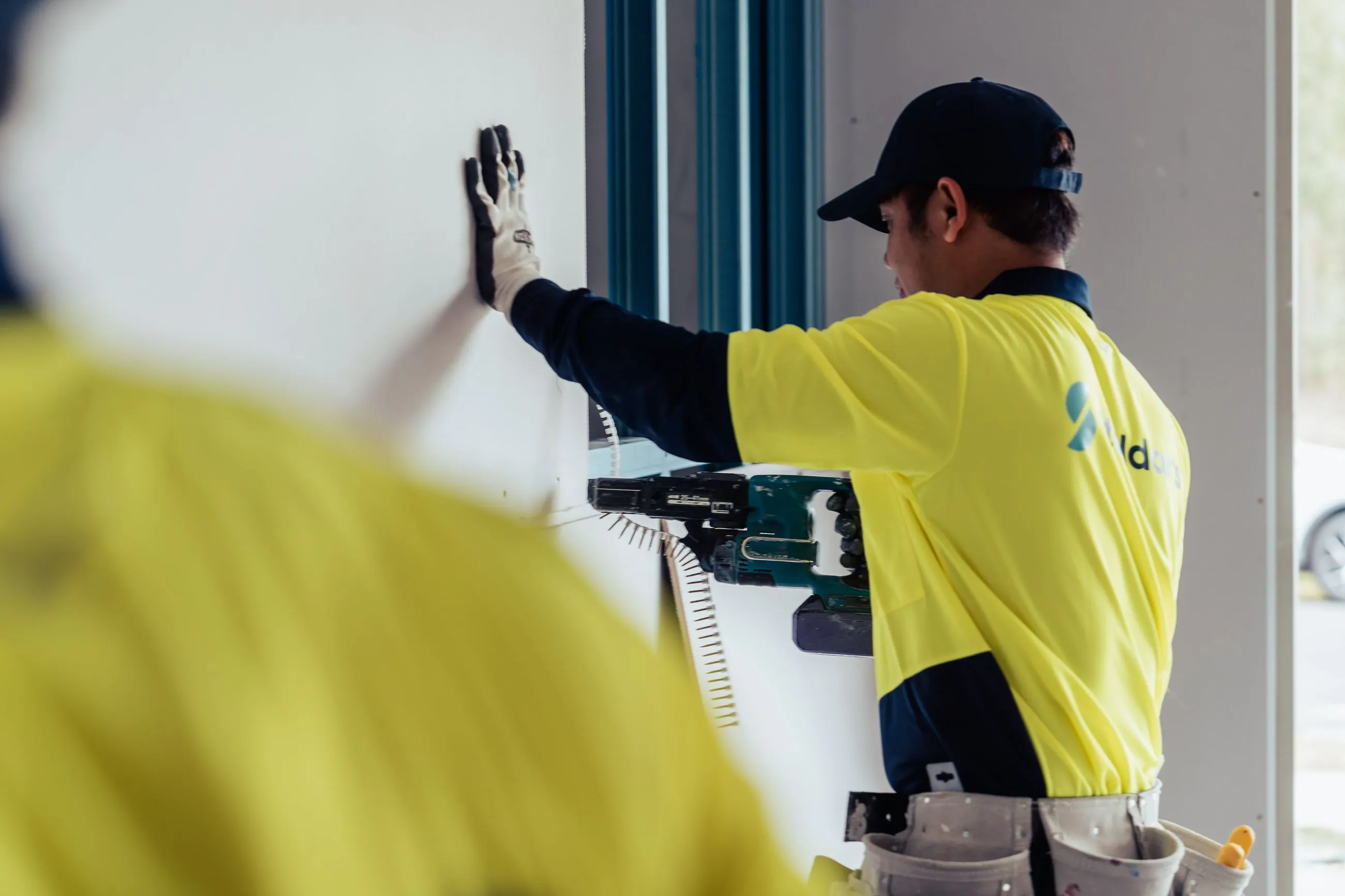 Worker in a yellow shirt and cap installing drywall using a power drill in an indoor construction site.