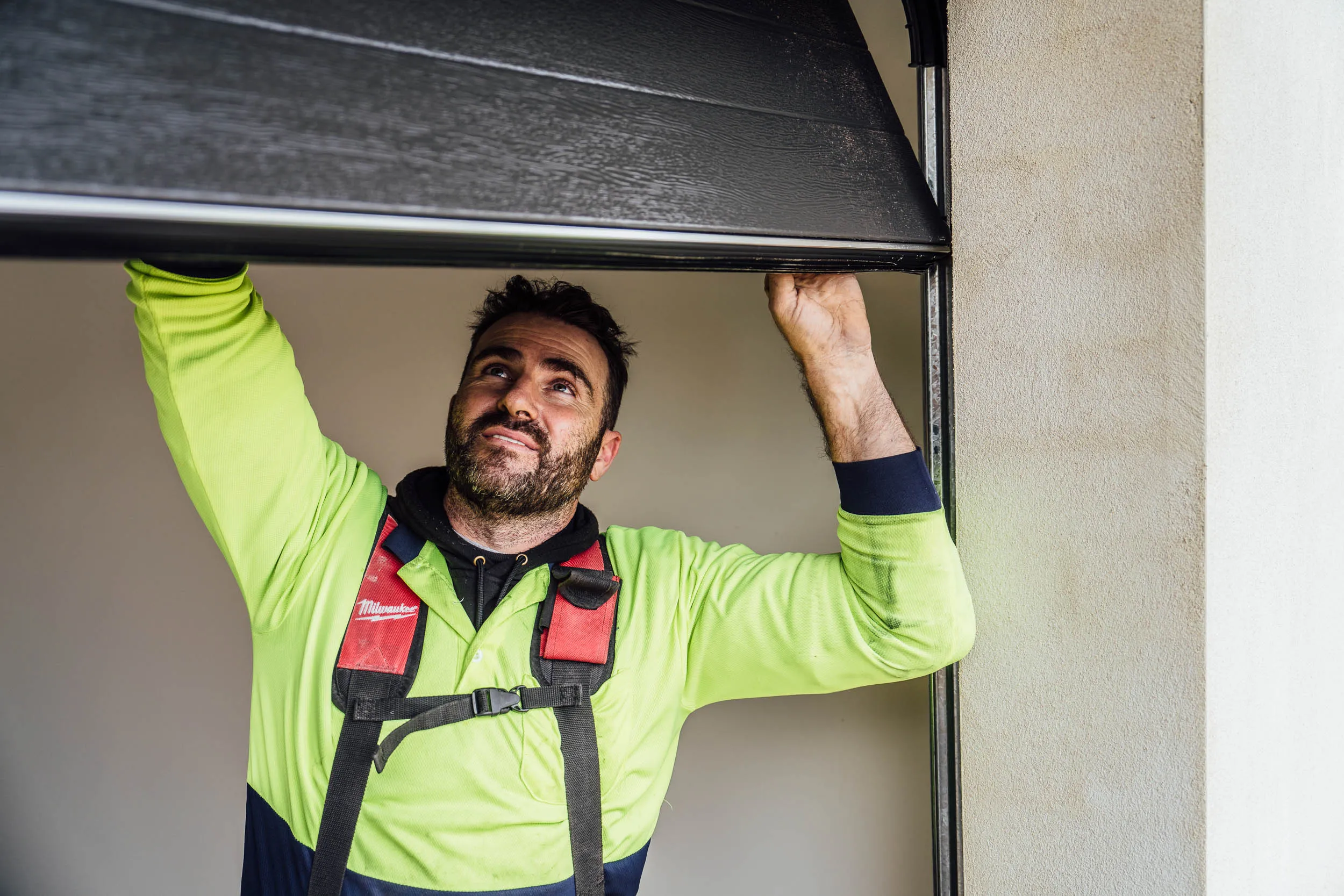 Man in safety harness inspecting or repairing a partially open black garage door indoors.