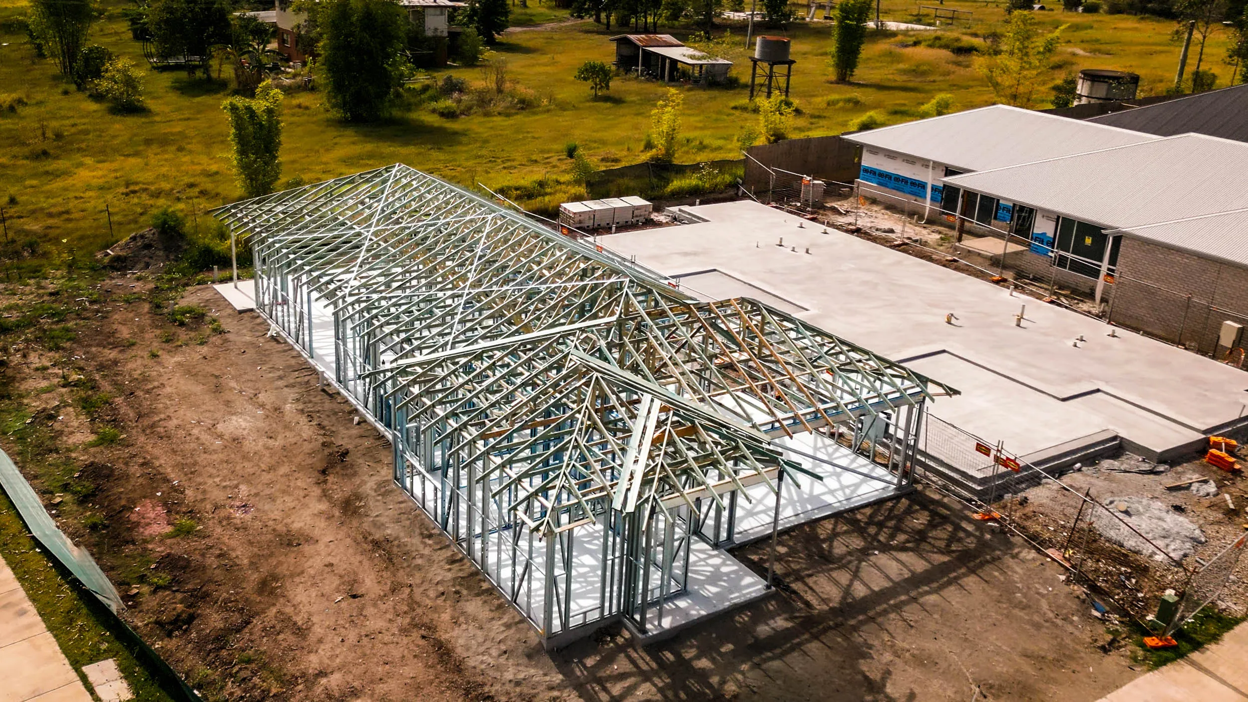 Metal framework of a building under construction on a cleared plot amid grassy area and nearby buildings.