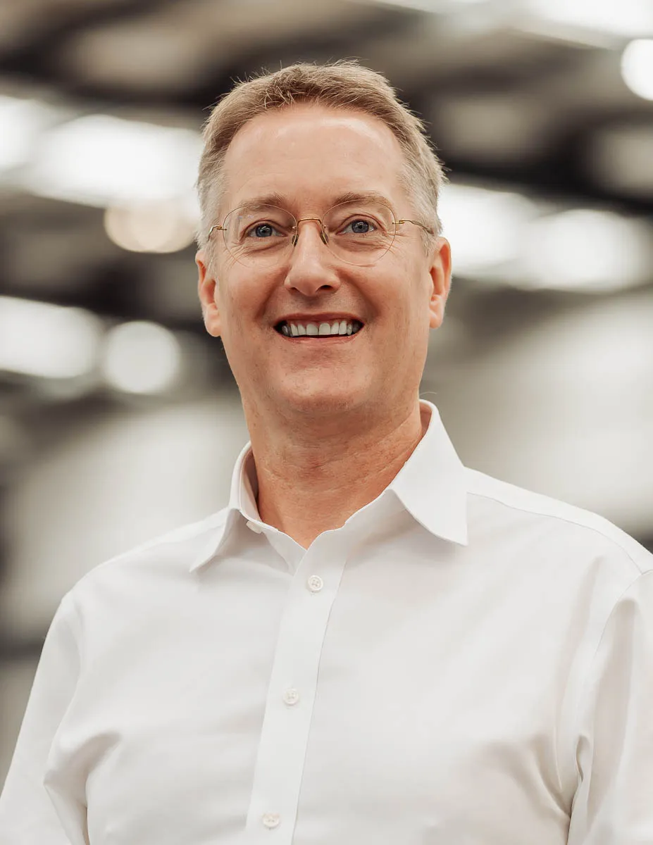 Smiling middle-aged man with glasses wearing a white button-down shirt against a blurred background.