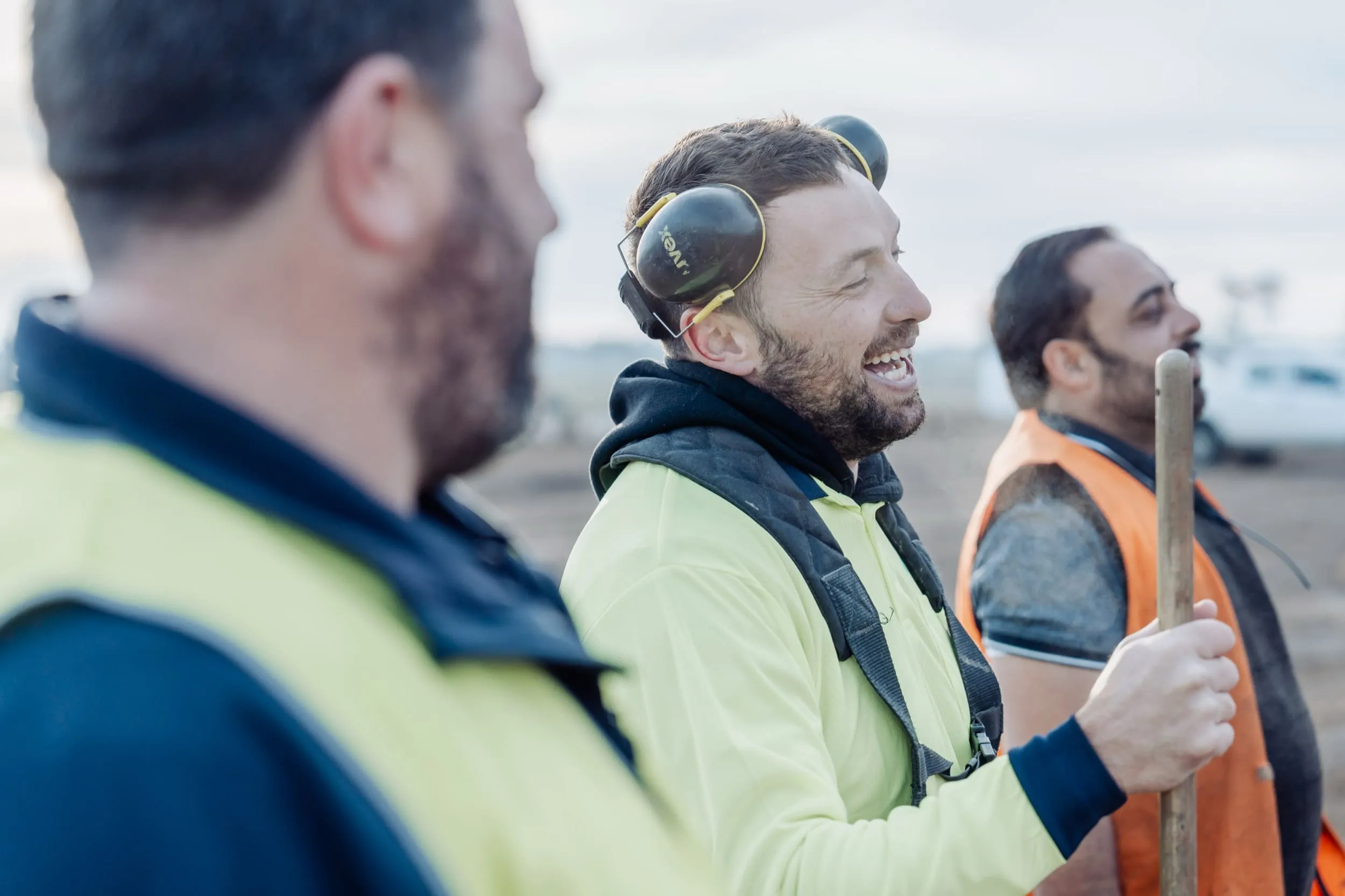 Three construction workers in high-visibility clothing laughing and talking outdoors at a work site.