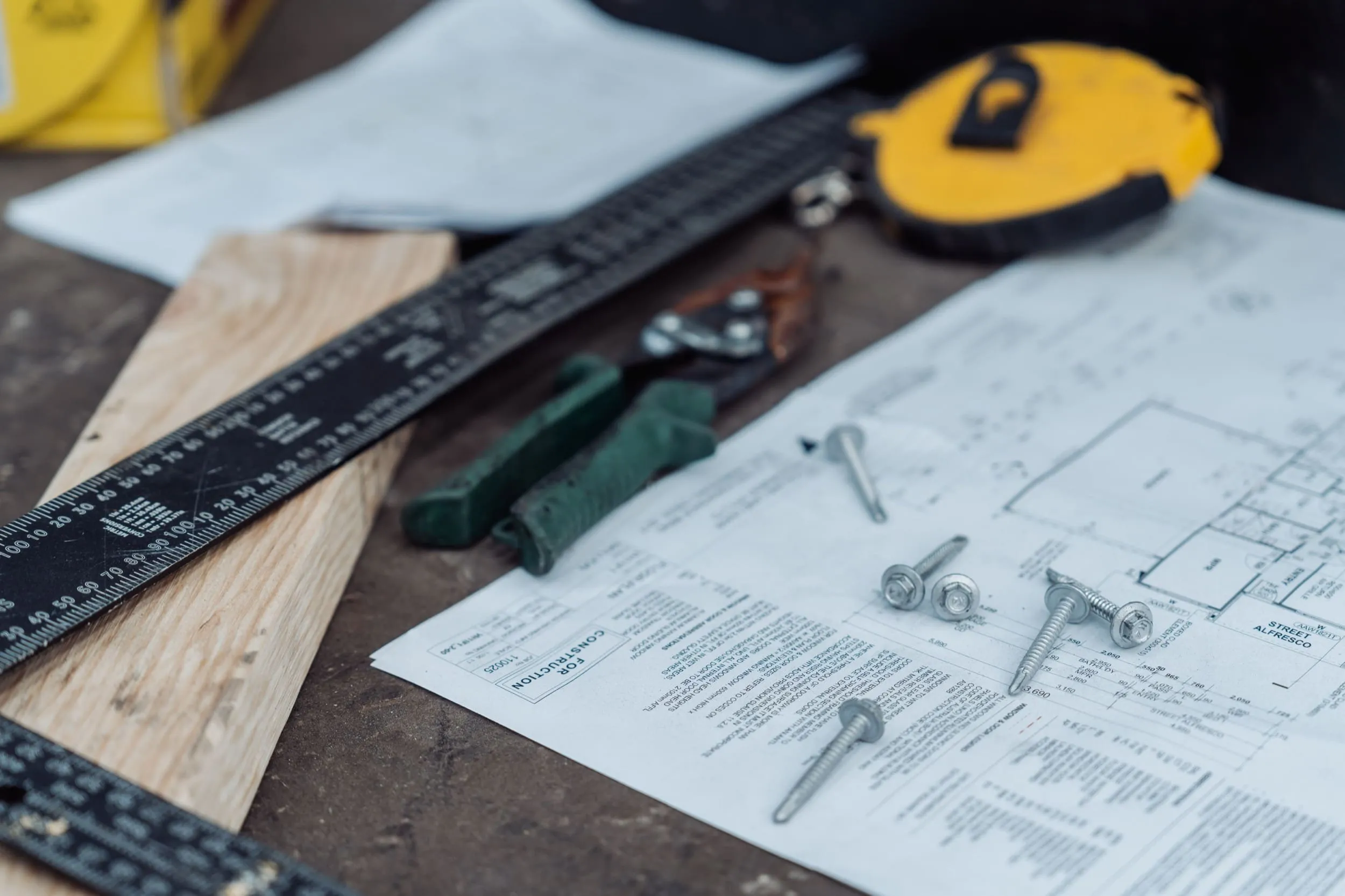 Construction plans with screws, a yellow tape measure, green handle tool, ruler, and wooden plank on a table.