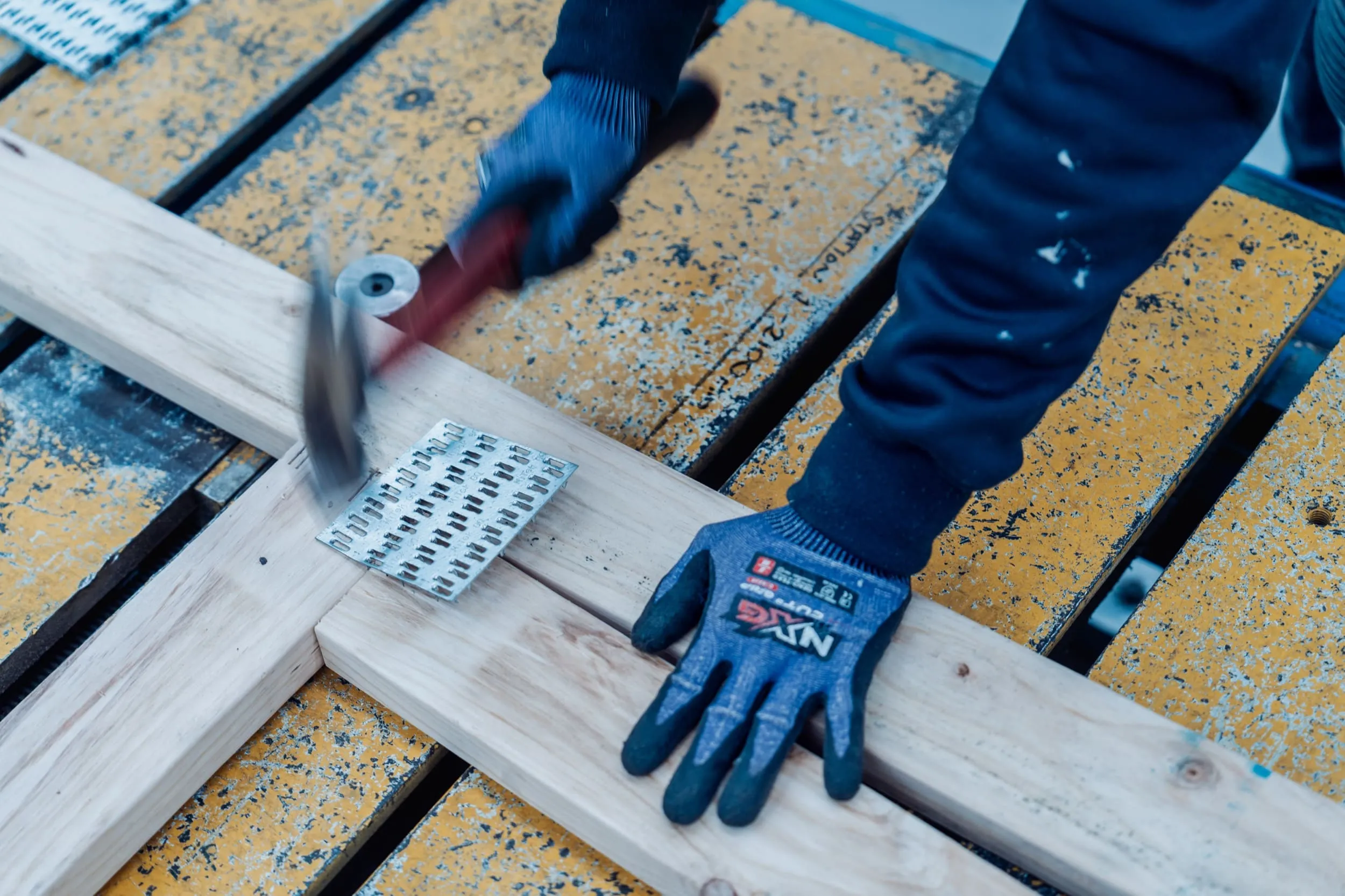 Person wearing gloves using a hammer to nail a metal connector on wooden planks over yellow surface.