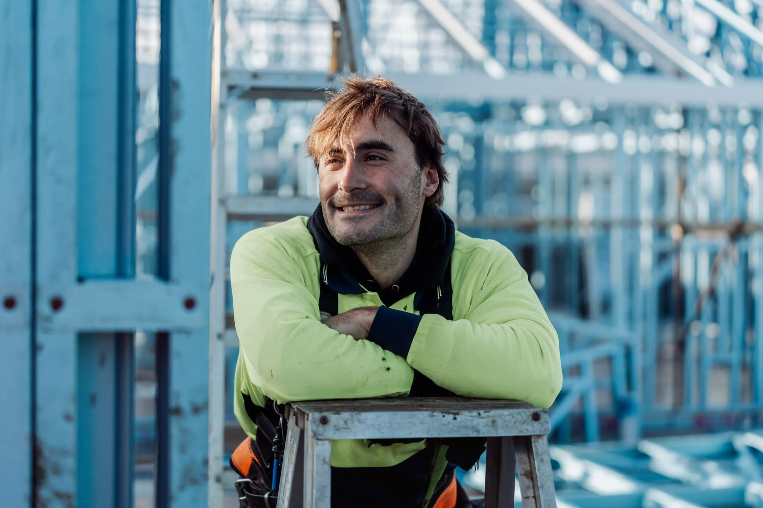 Smiling construction worker in a yellow jacket leaning on a metal ladder at a worksite.