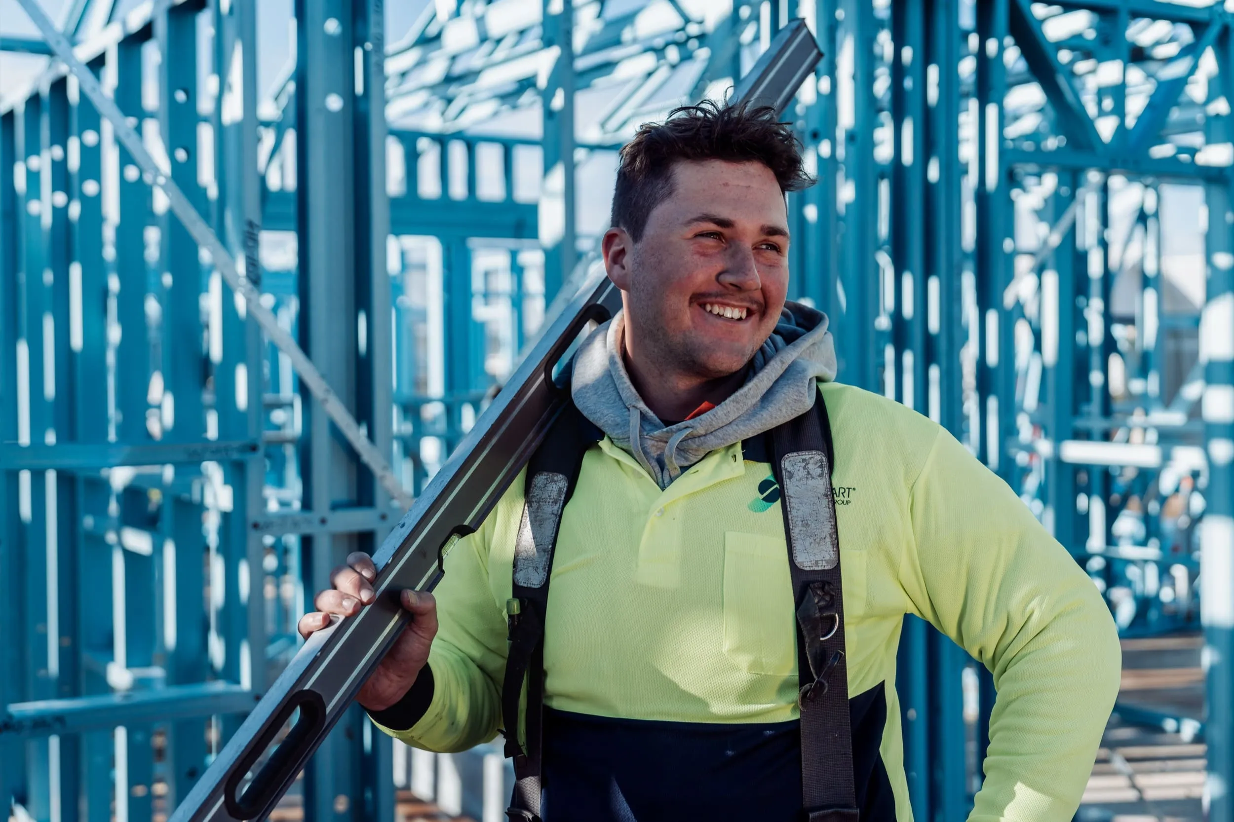 Smiling construction worker in yellow high-visibility shirt holding a level on his shoulder at a steel framework site.