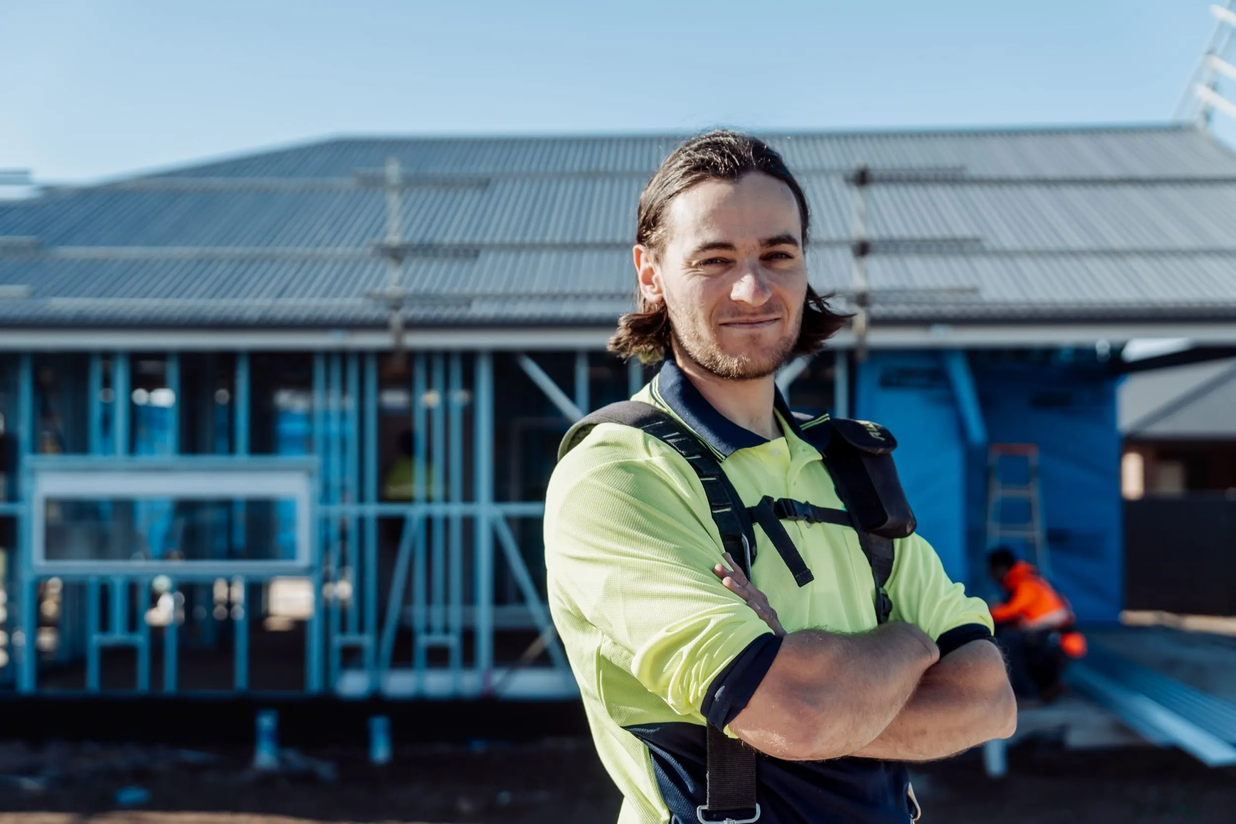 Confident construction worker with long hair in yellow high-visibility shirt stands with arms crossed at site.