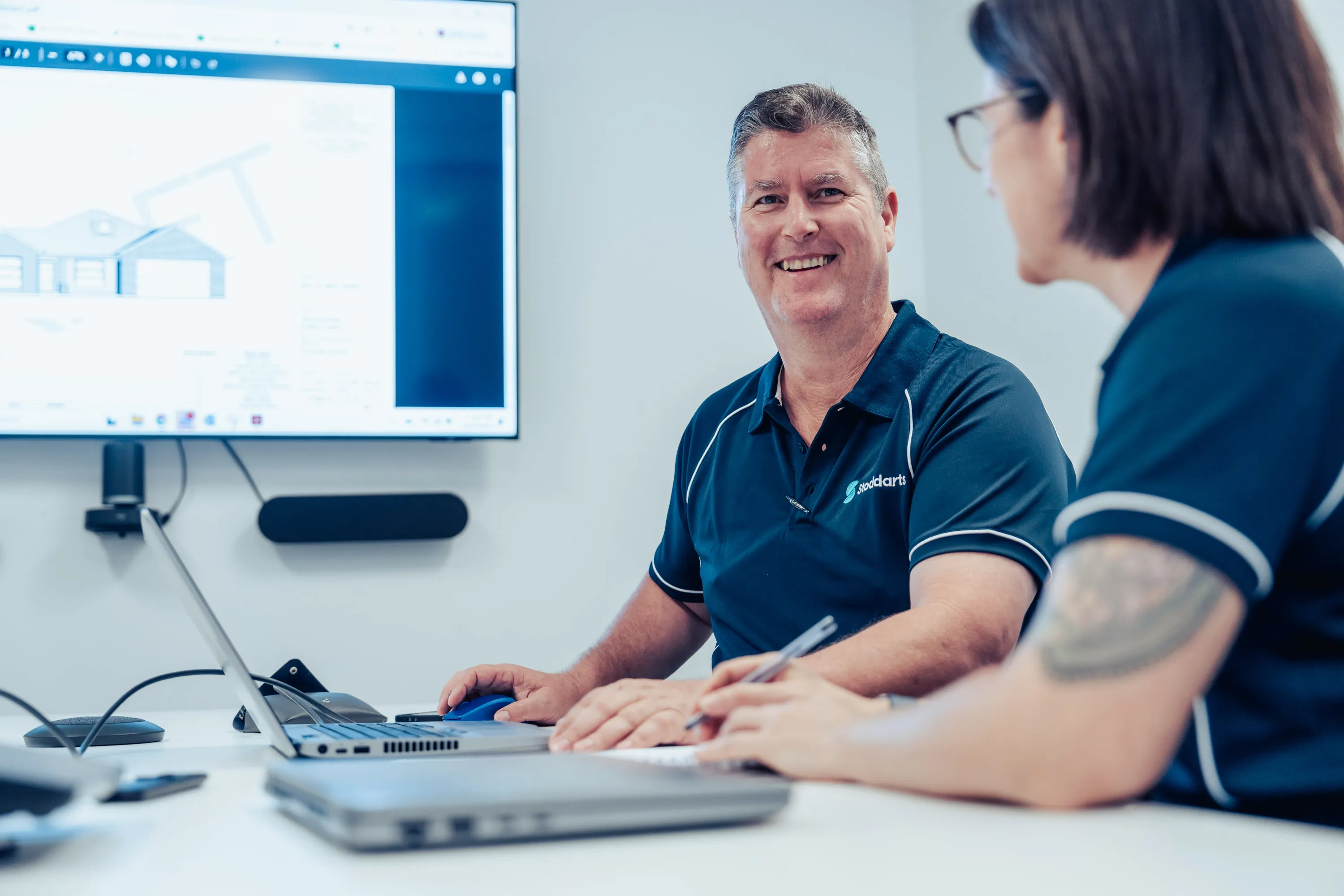 Two colleagues in navy shirts discussing house plans on a laptop in a conference room.