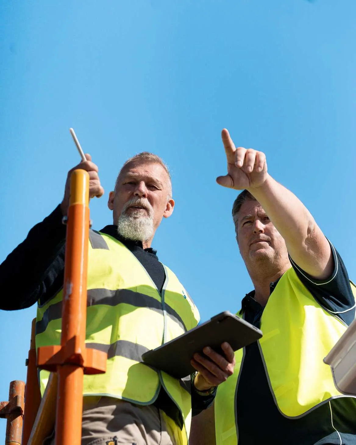 Two male construction workers in yellow vests, one holding a tablet, the other pointing up against blue sky.