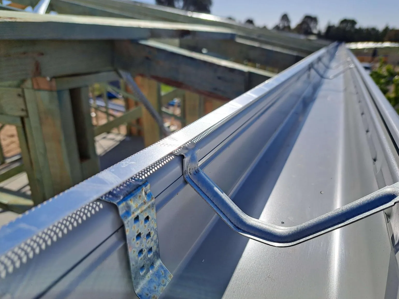 Close-up of a metal rain gutter attached to a wooden roof frame under construction on a sunny day.