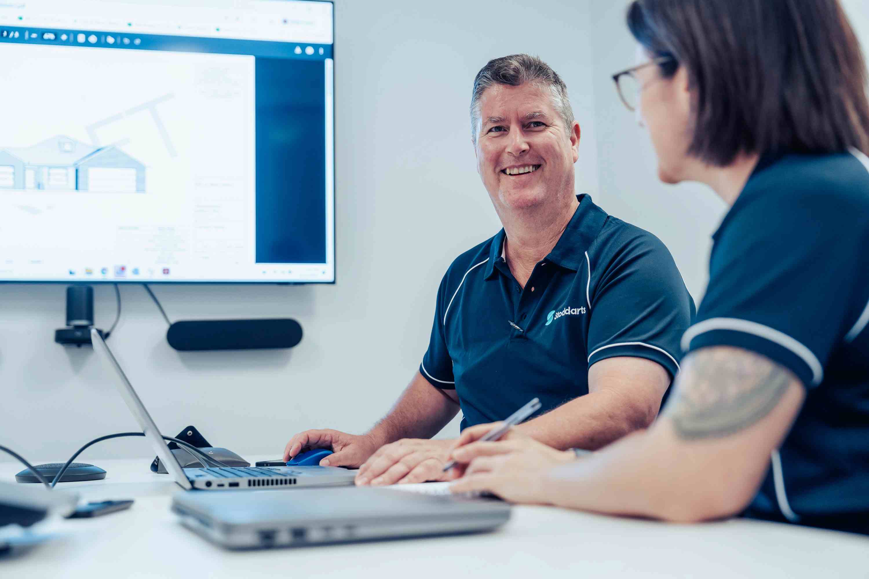 Two people in matching navy shirts discussing plans with a laptop and blueprint on screen.