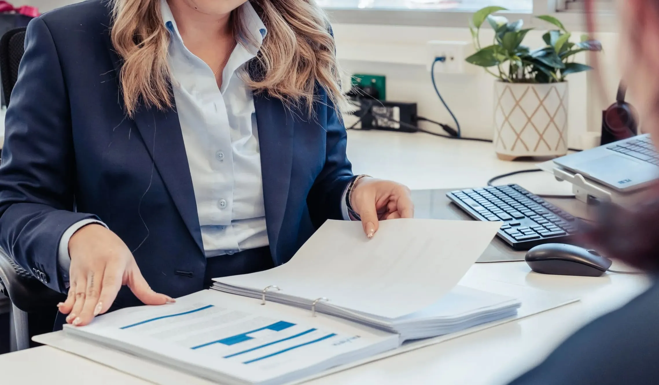 Businessperson in a suit reviewing a binder with charts at an office desk with keyboard and plant.