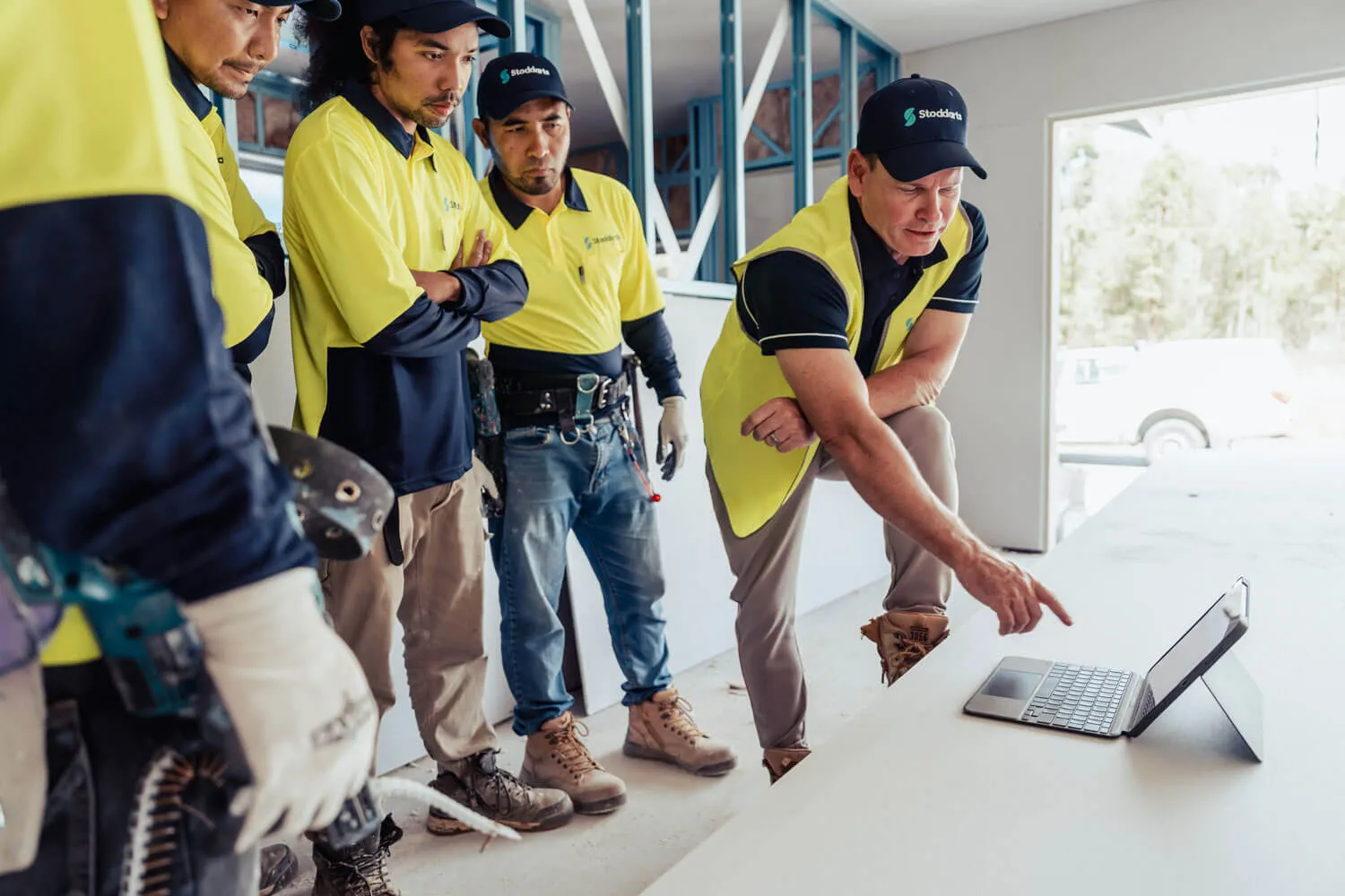 Construction workers in yellow and black uniforms attentively watching a man pointing at a tablet screen.
