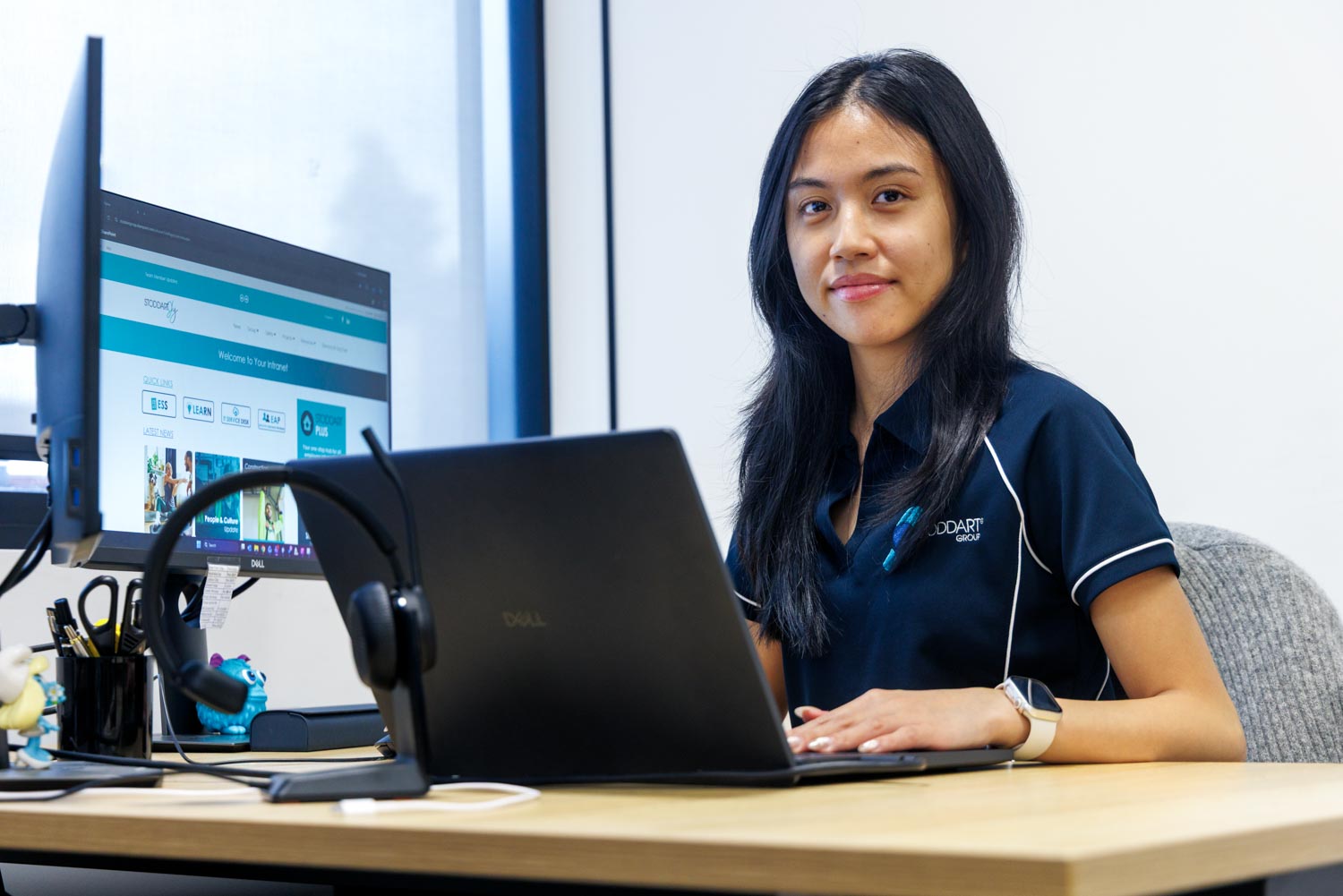 Woman in navy polo working on a Dell laptop at a desk with a headset and desktop monitor.