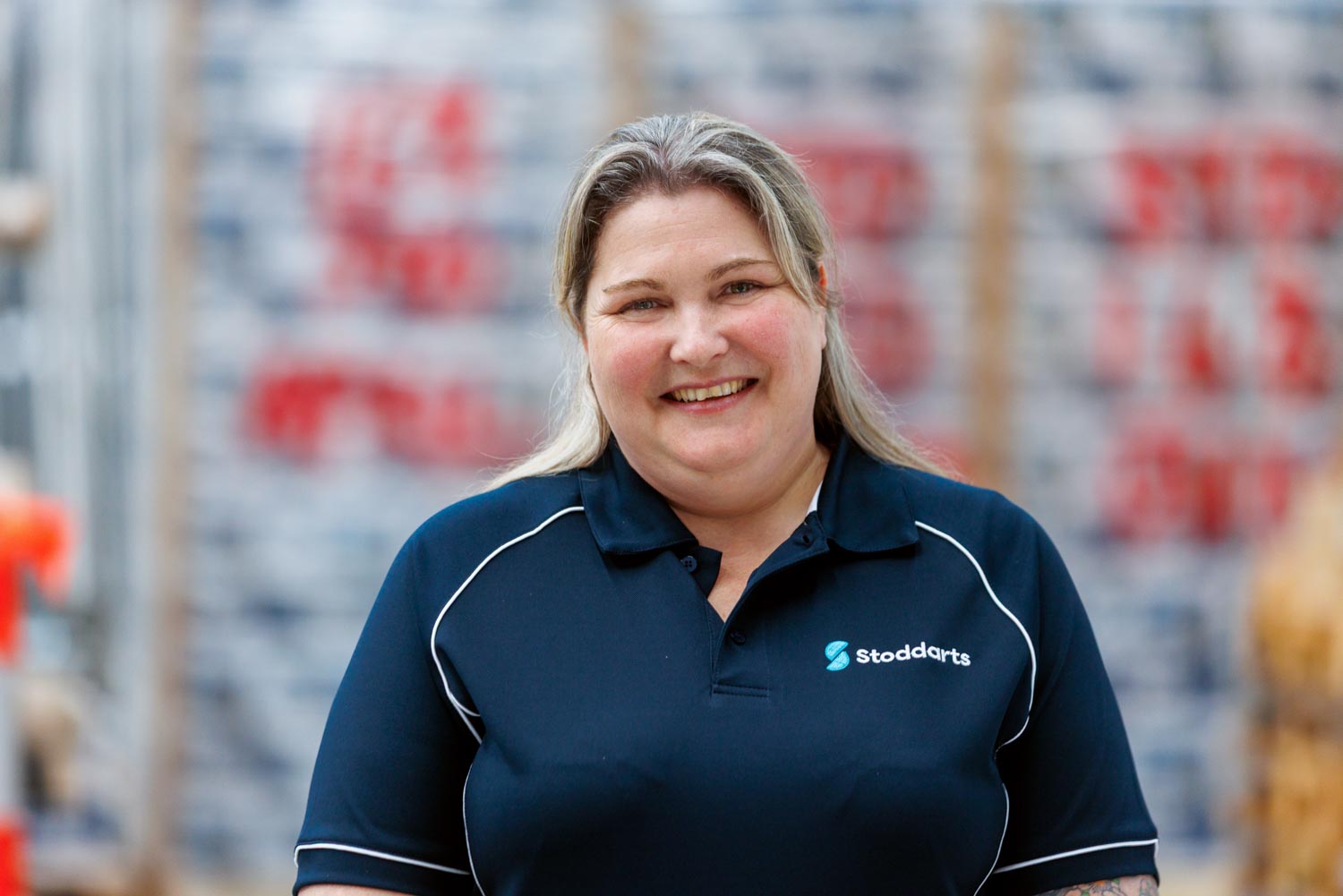 Smiling woman in a navy Stoddarts polo shirt standing in a warehouse with pallet stacks behind her.