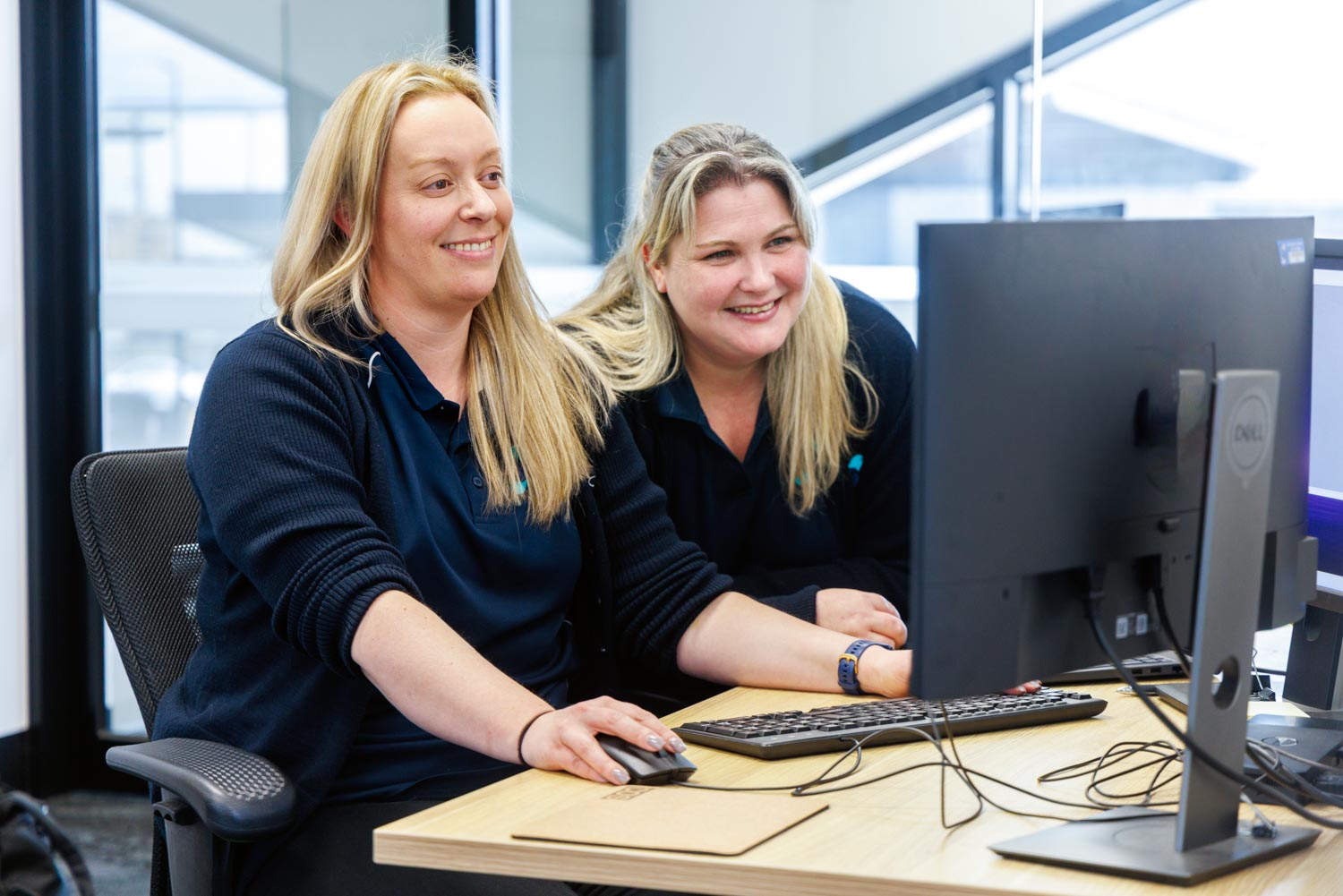 Two women smiling and working together at a desk with a desktop computer in a bright office.